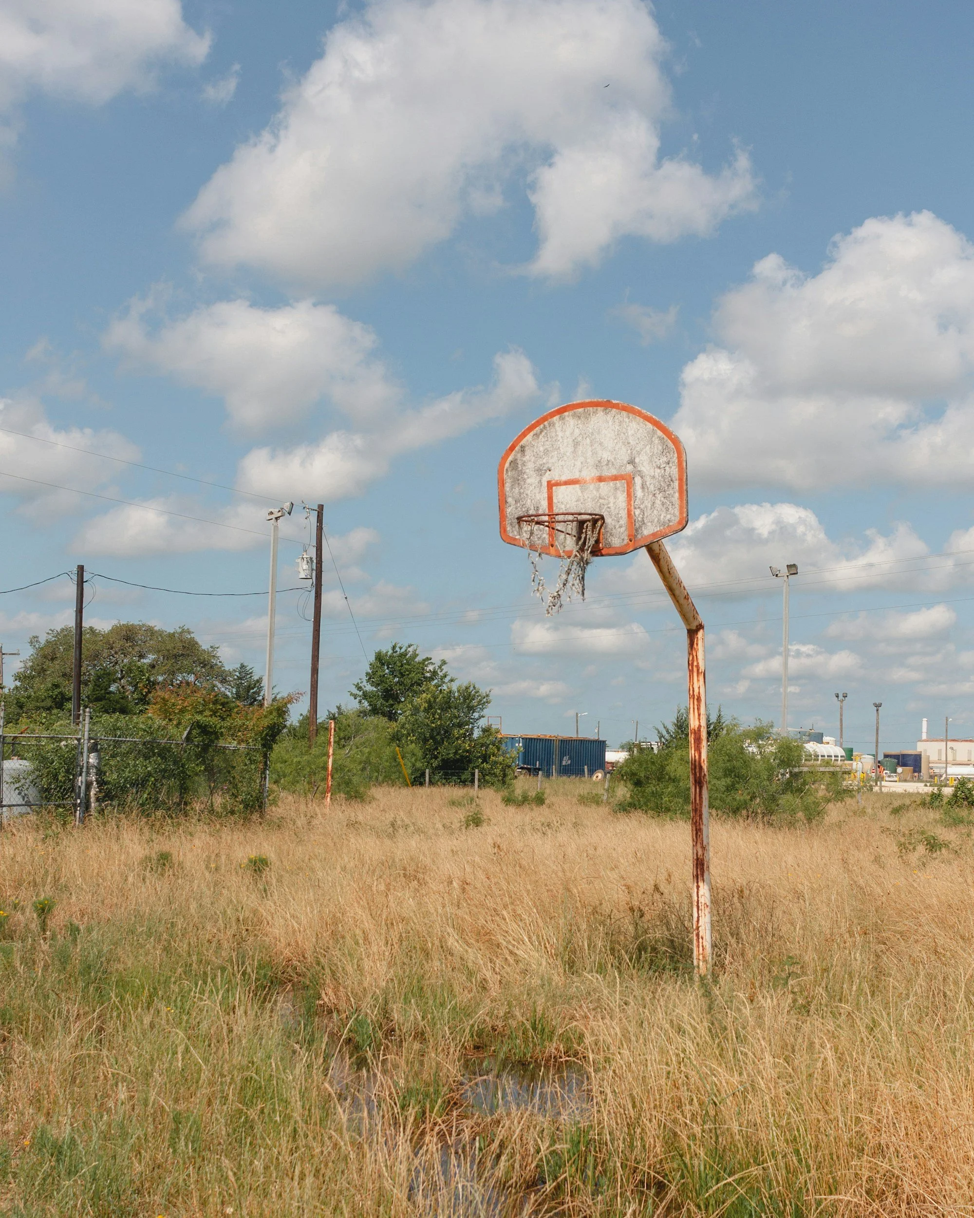 An old, rusted basketball hoop with a worn net is situated in a grassy field. The sky above is mostly clear with a few scattered clouds, and there are power lines and trees in the background.