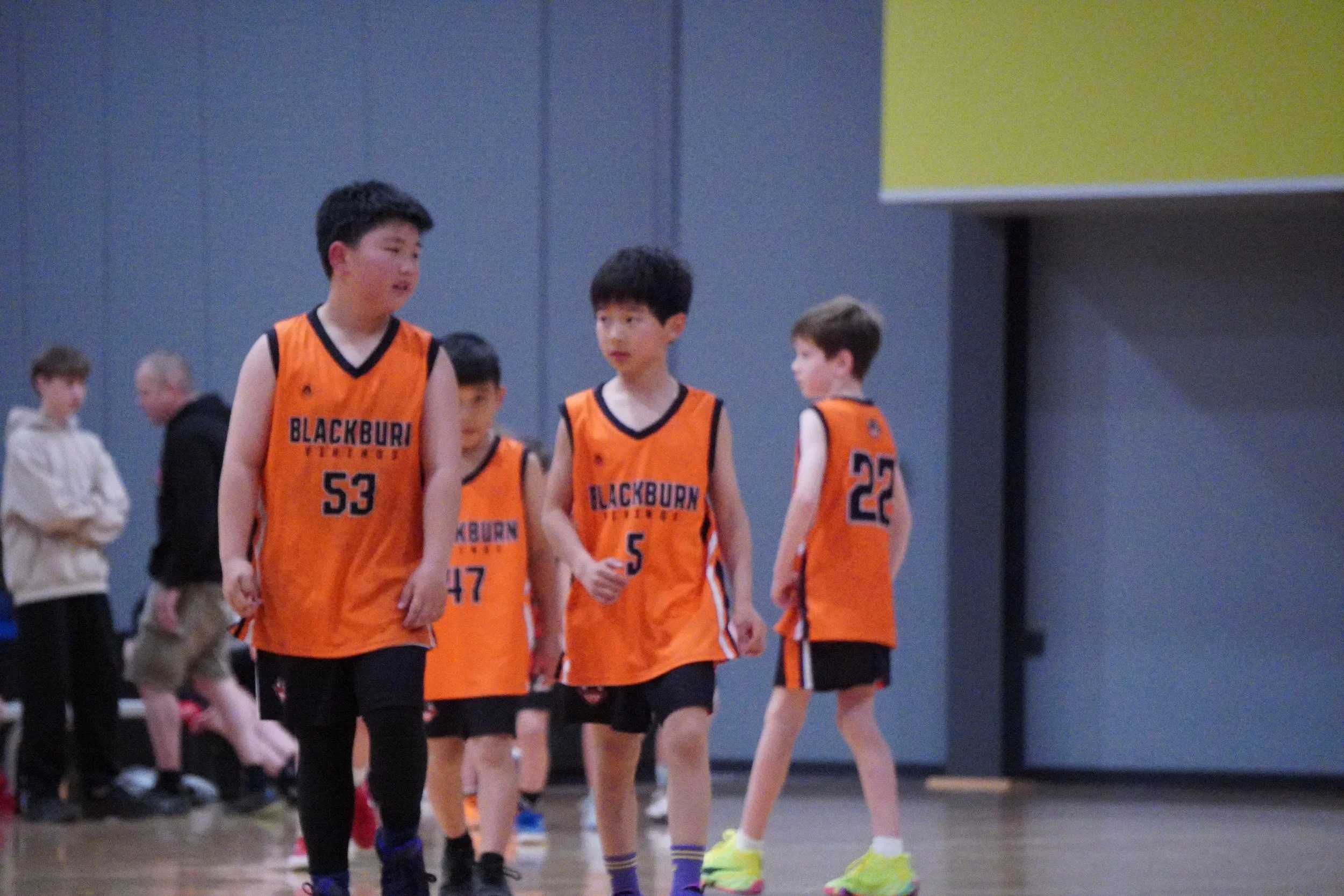 A group of young boys playing basketball indoors, wearing orange jerseys with black and white accents and the logo 'Blackburn' on the front. Some boys are walking, while others are standing, with a plain indoor gym background and a few adults in the distance.