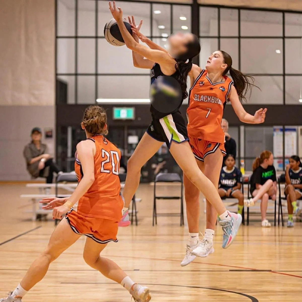 Young female basketball players in orange and black uniforms competing during a game, with one player jumping to block a shot and others watching.