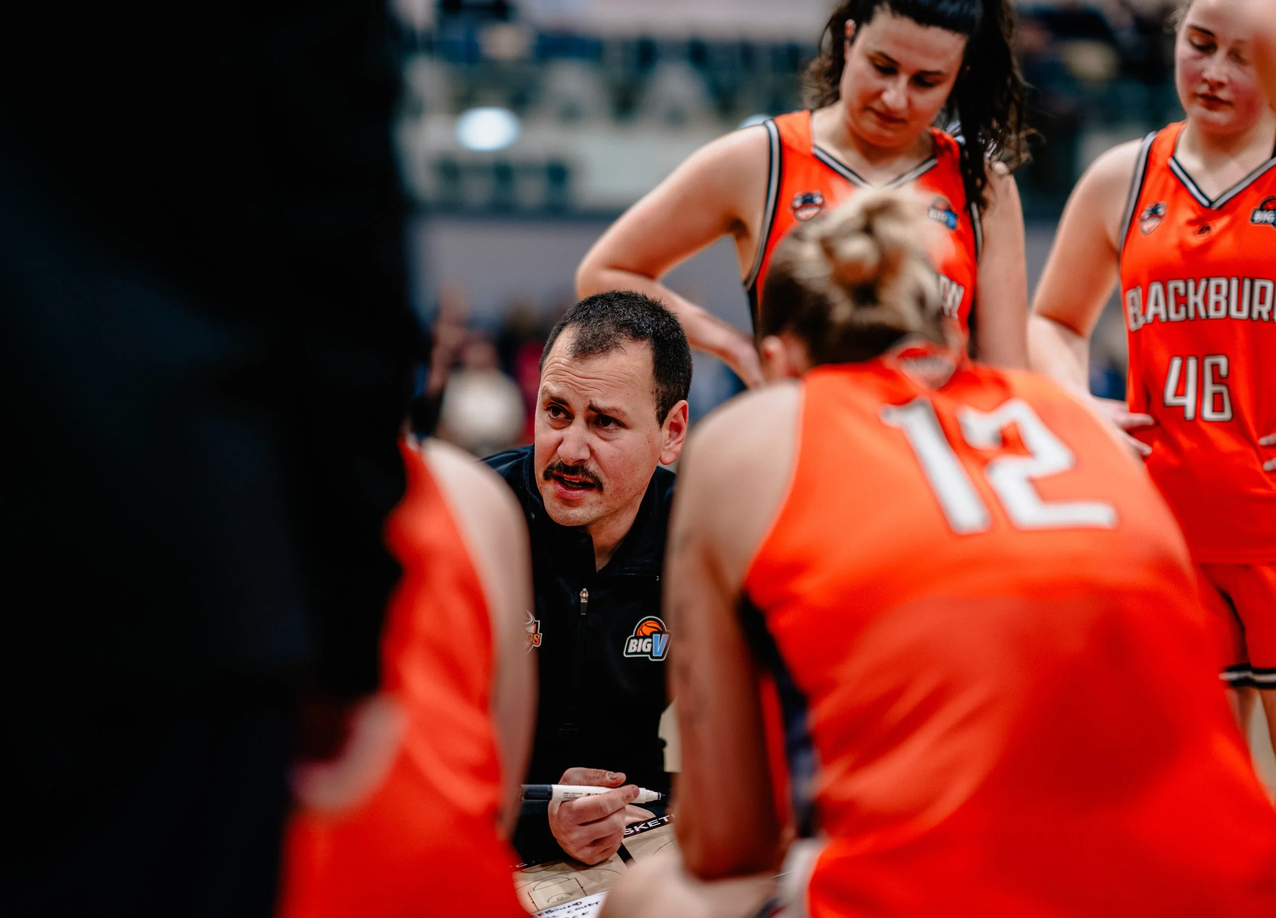 A women's basketball team in orange jerseys during a timeout huddle, with their coach speaking to the players.