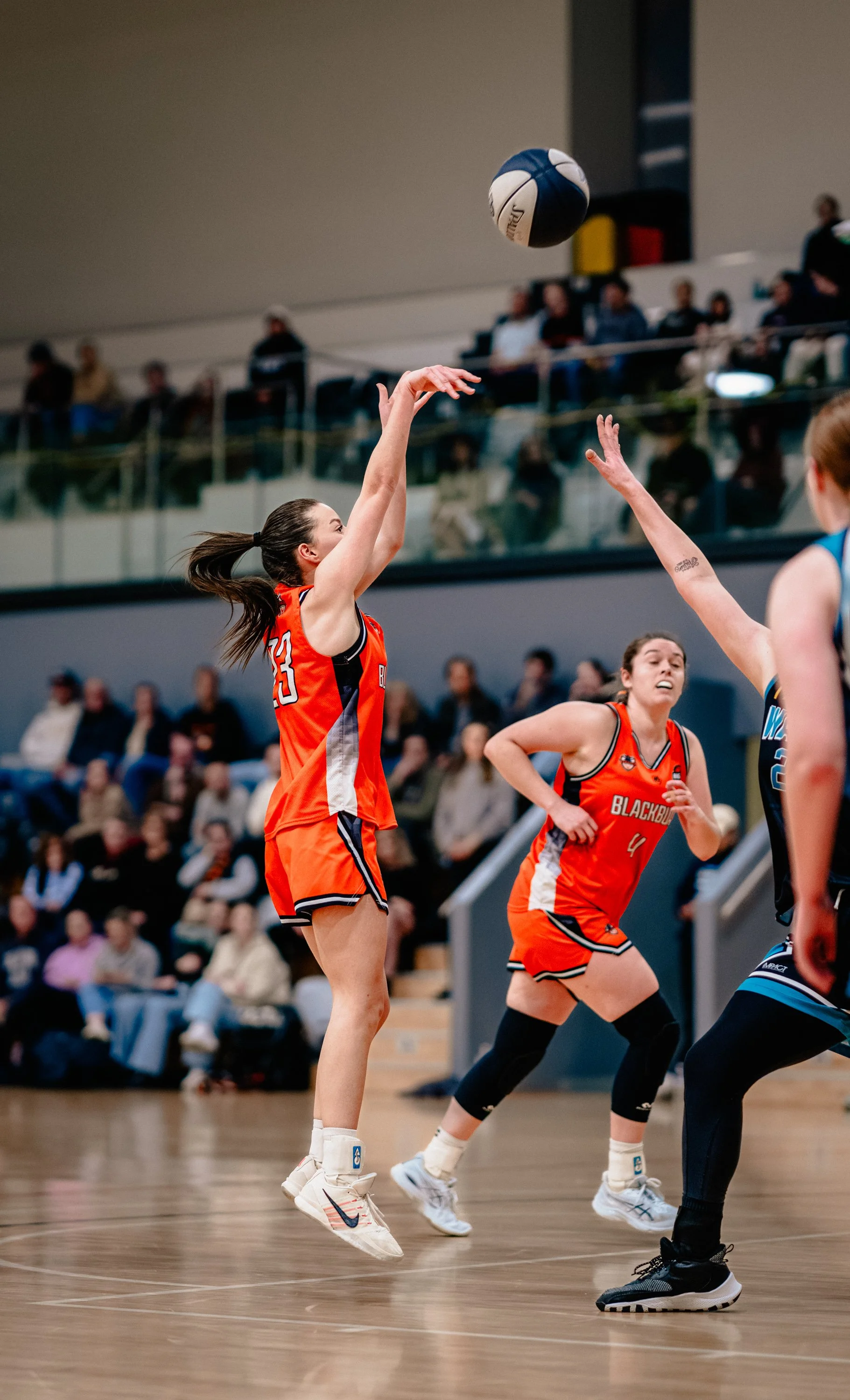 A women's basketball game in progress with players jumping to catch the ball, players in orange jerseys and a crowd watching in the background.