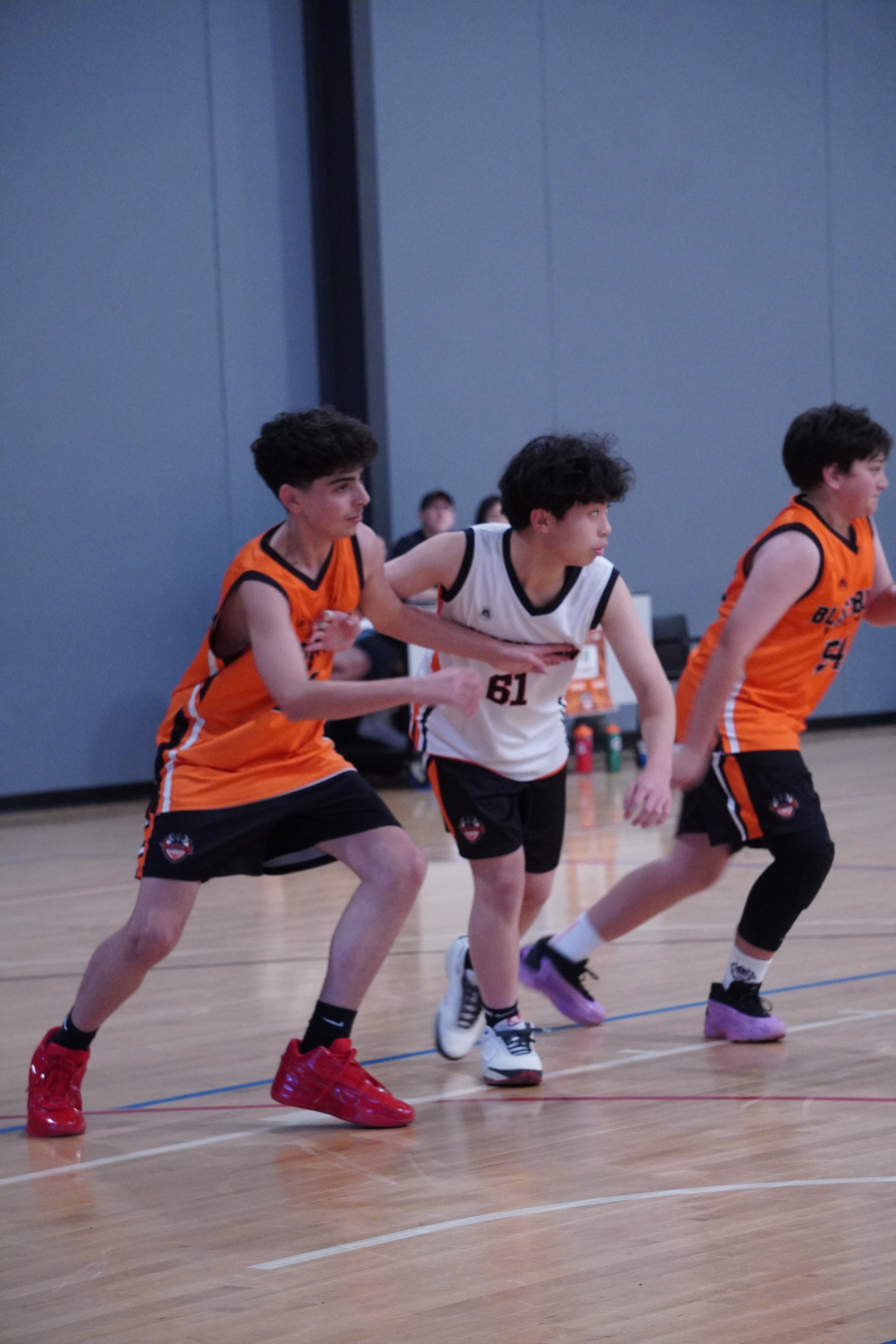 Three young male basketball players in orange and white jerseys running on the court during a game.