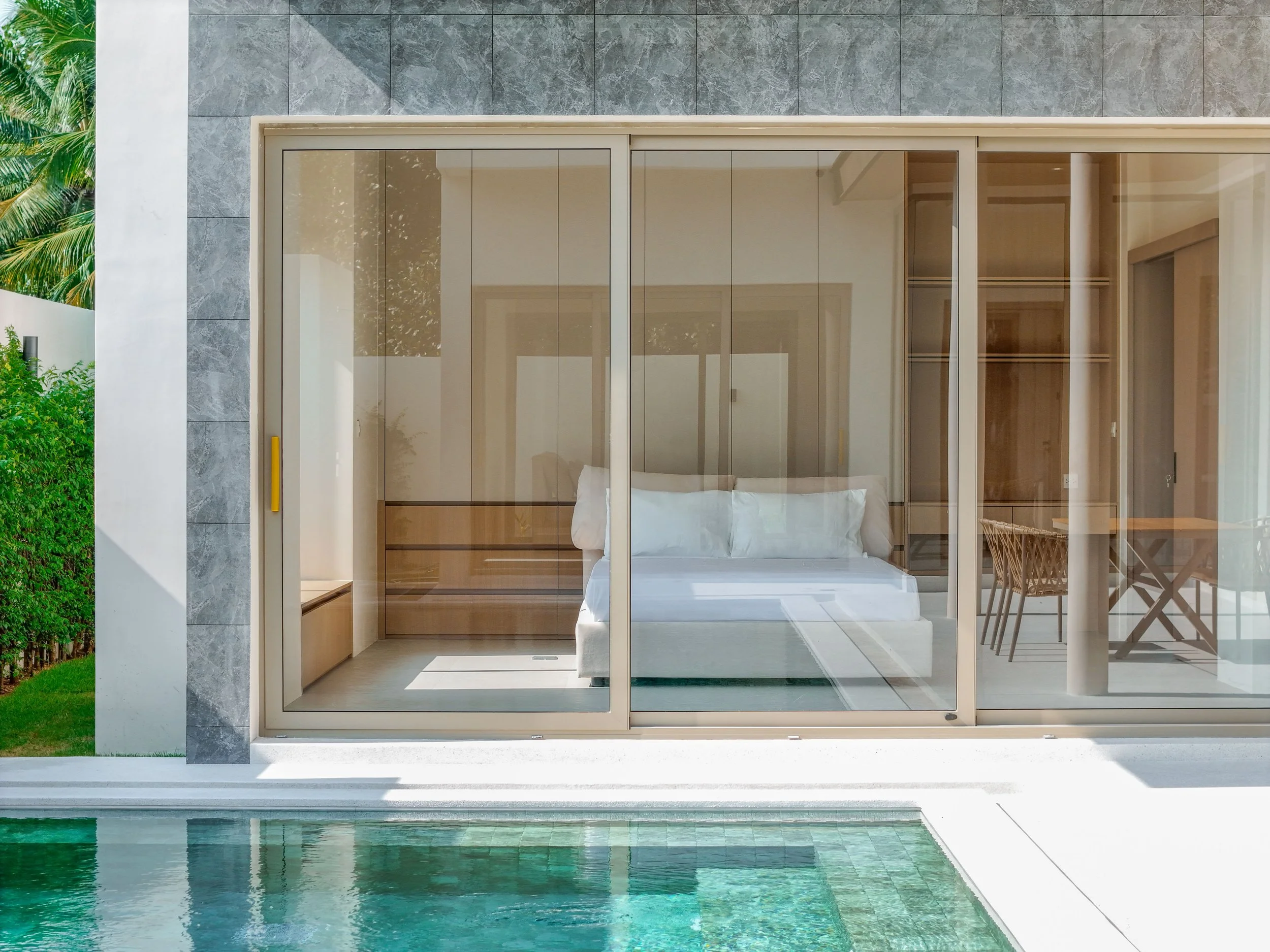 Das Bild zeigt ein modernes Schlafzimmer, das durch große Glasfenster vom angrenzenden Poolbereich getrennt ist. Das Zimmer ist minimalistisch eingerichtet, mit einem weißen Bett und einem offenen Kleiderschrank aus Holz im Hintergrund. Durch die Fen