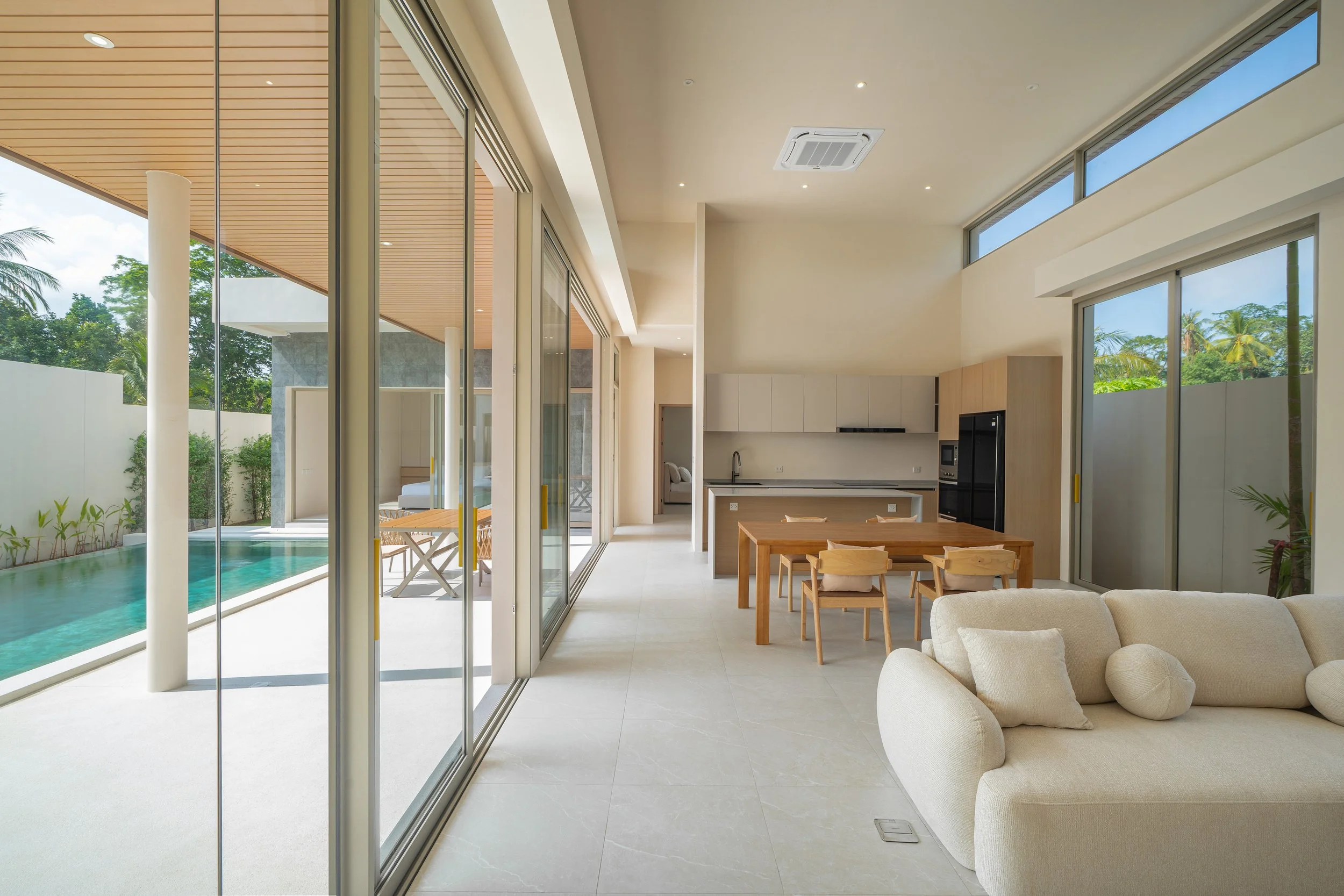 Innenraum eines modernen, offenen Wohnbereichs mit großen Glasfenstern, Blick auf einen Pool im Außenbereich, eine weiße Couch, ein Holztisch mit Stühlen, weiße Wände und eine Küche im Hintergrund.