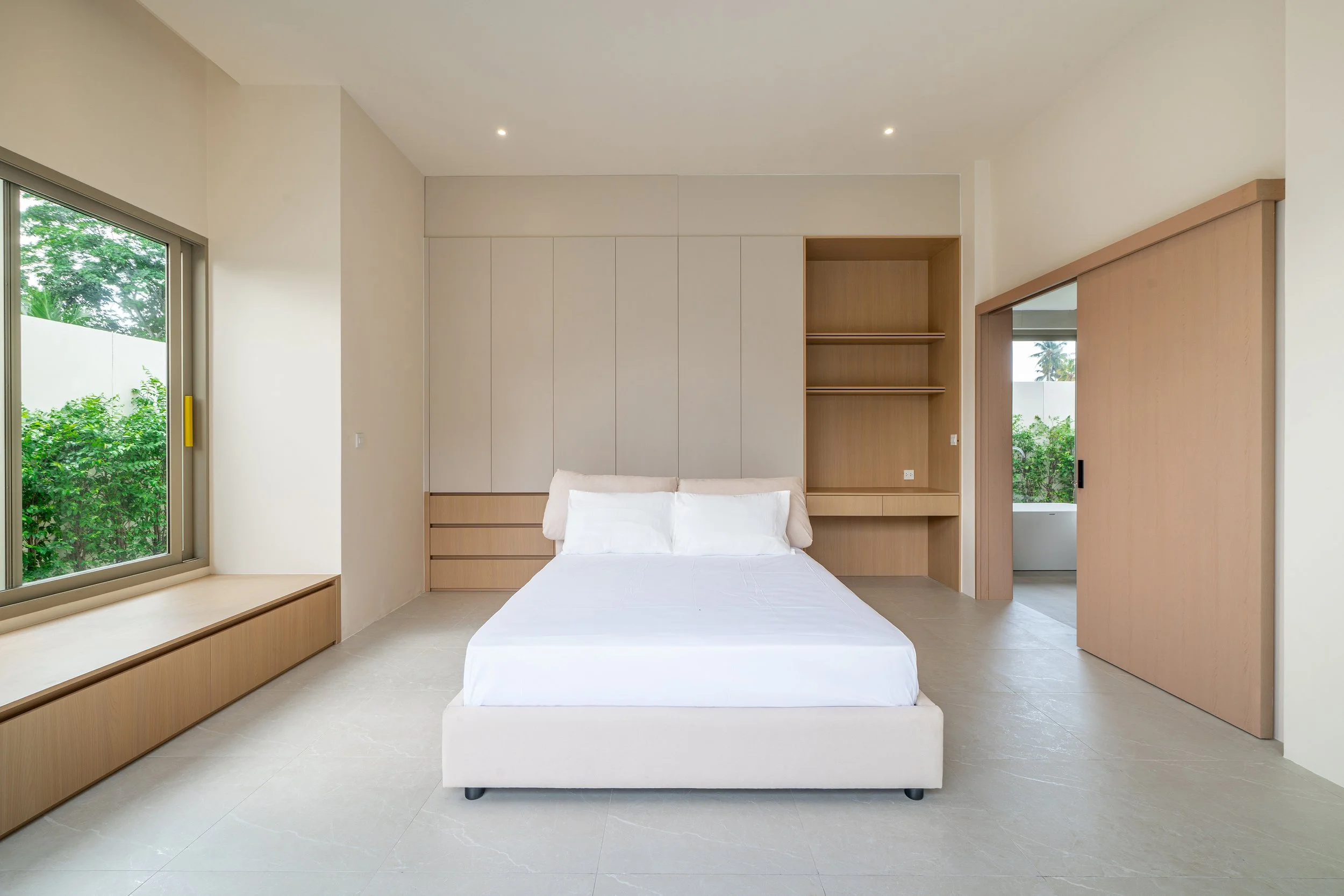 Ein modernes Schlafzimmer mit einem weißen Bett, großen fensters und viel Stauraum in hellen Holztönen.