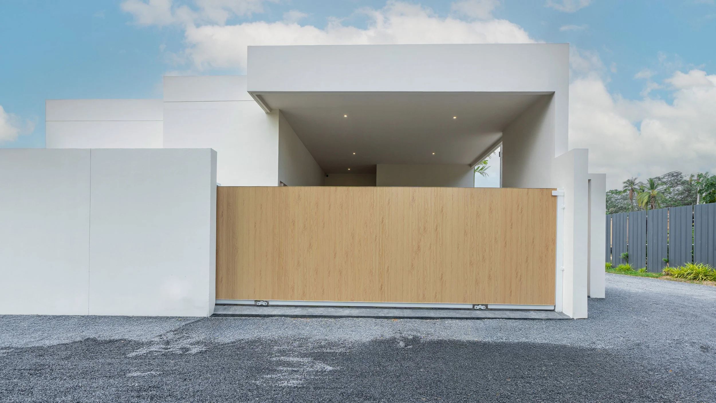 Modernes, weißes Haus mit Holztor vor grauem Boden und blauer Himmel mit Wolken.