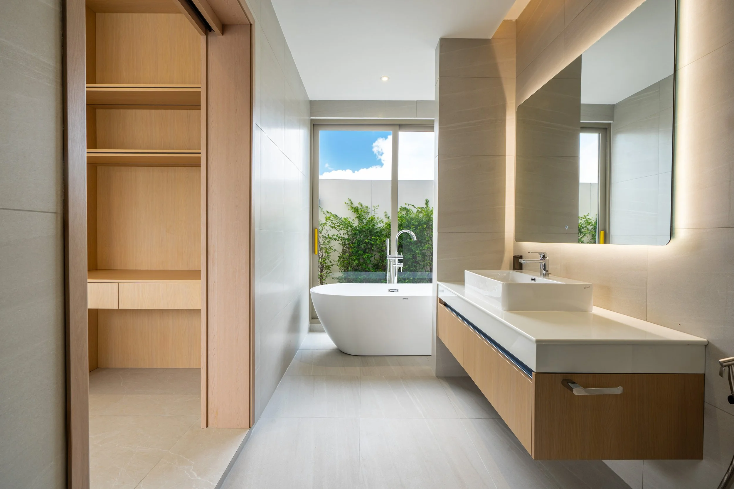 Modernes Badezimmer mit freistehender Badewanne, Waschtisch mit Spiegel und einem großen Fenster mit Blick nach draußen auf grünes Gebüsch und Himmel.