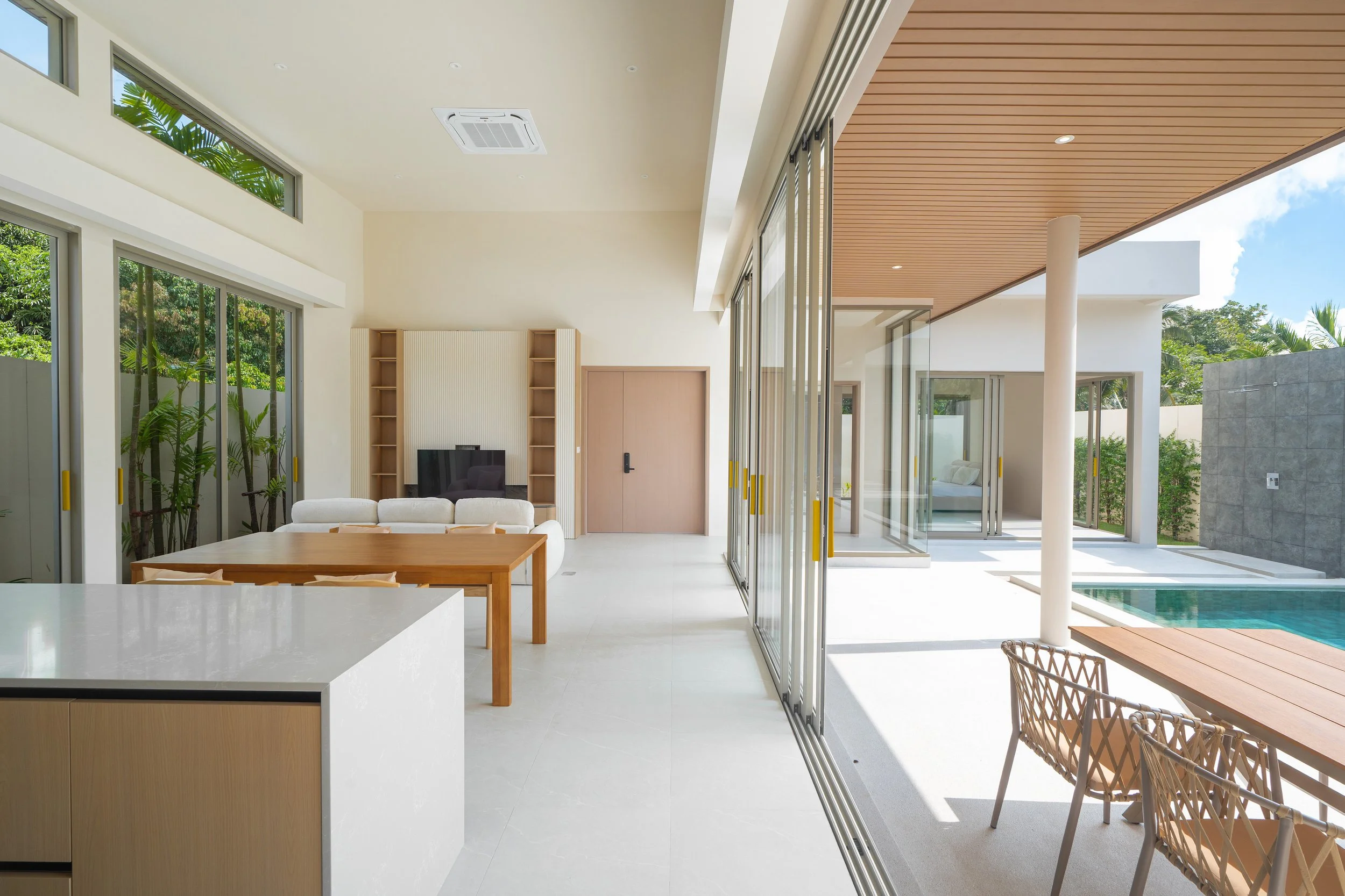 Innenansicht eines modernen Hauses mit großen Glastüren, einem Wohnzimmer, einem Essbereich und einem Außenbereich mit Pool, Sonnenlicht und grünem Garten.