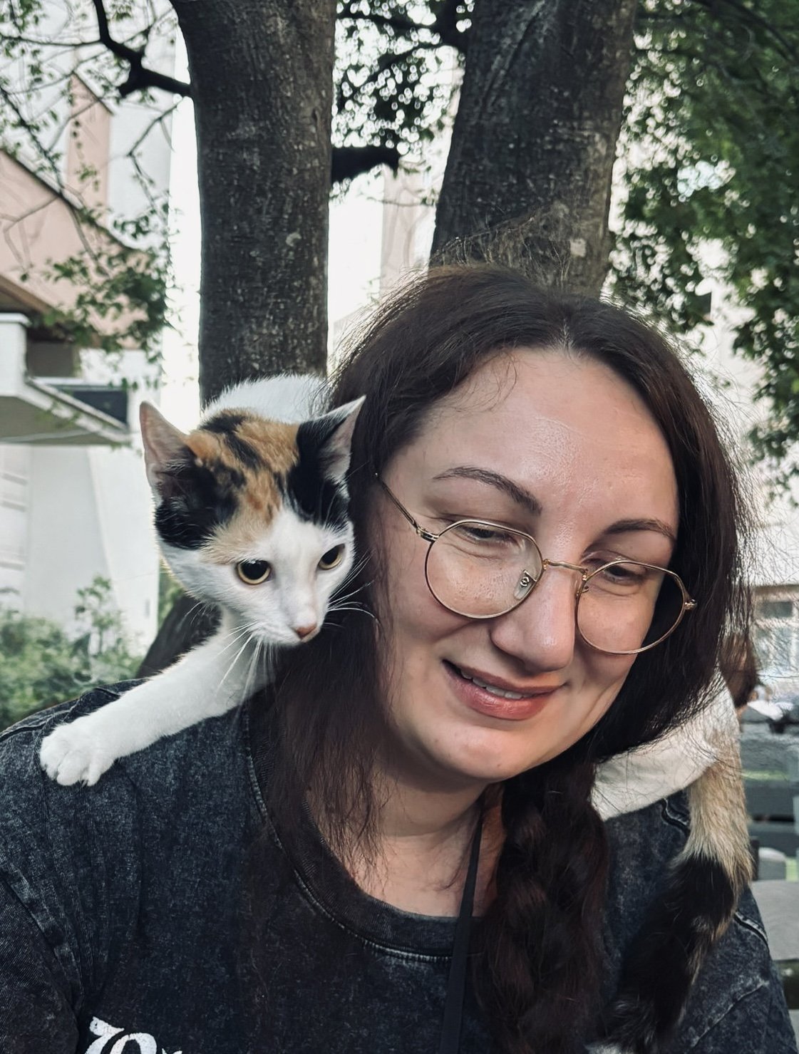 A woman with glasses and long dark hair smiling while a calico cat with white, orange, and black fur rests on her shoulder. They are outdoors with trees and buildings in the background.