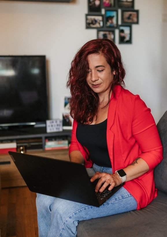 A woman with curly red hair sitting on a couch and working on a laptop, wearing a red blazer, black top, and a smartwatch, in a living room with a TV and photo frames on the wall.