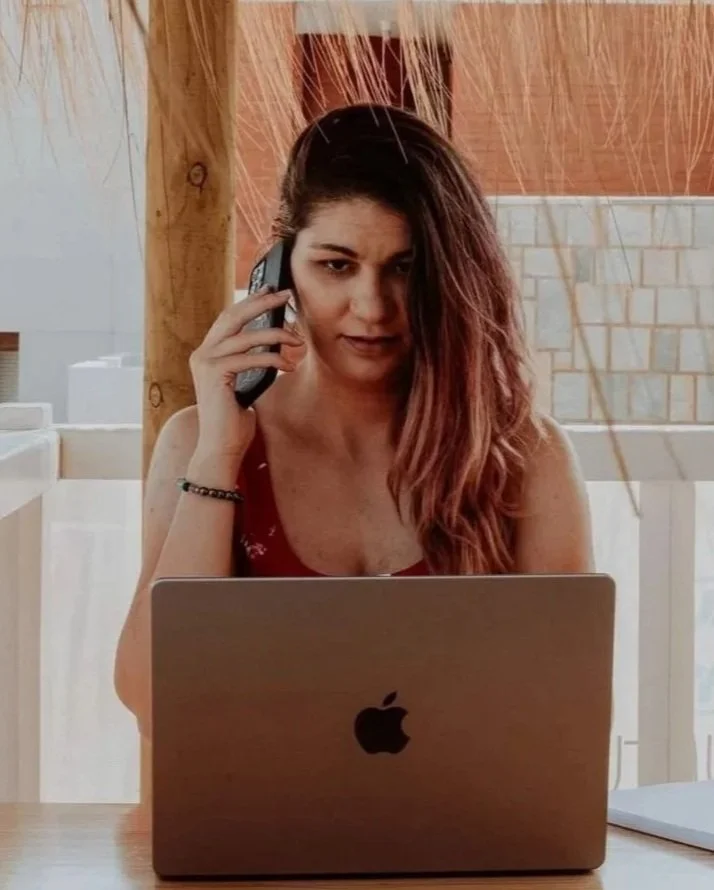 A woman with long brown hair holding a phone to her ear while sitting at a table with an open MacBook in front of her.