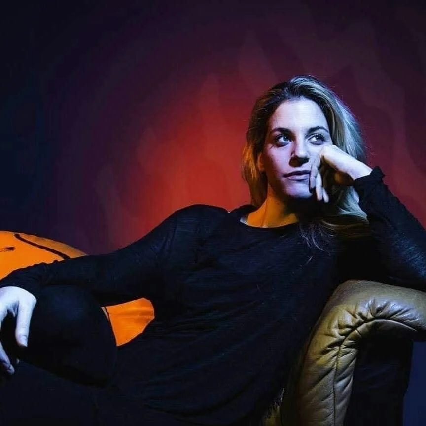 Woman in black top sitting on a tan leather chair, with her chin resting on her hand, looking thoughtfully to the side, against a dark background with red and purple lighting.