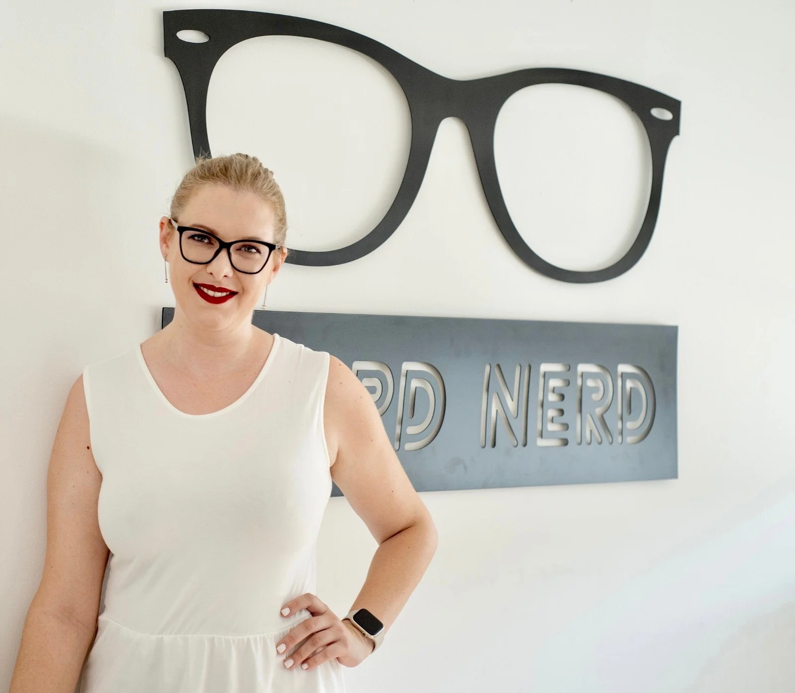 A woman with blonde hair and glasses smiling, standing in front of a wall with a large black eyeglasses illustration and a sign that says "Nerd Nerd".