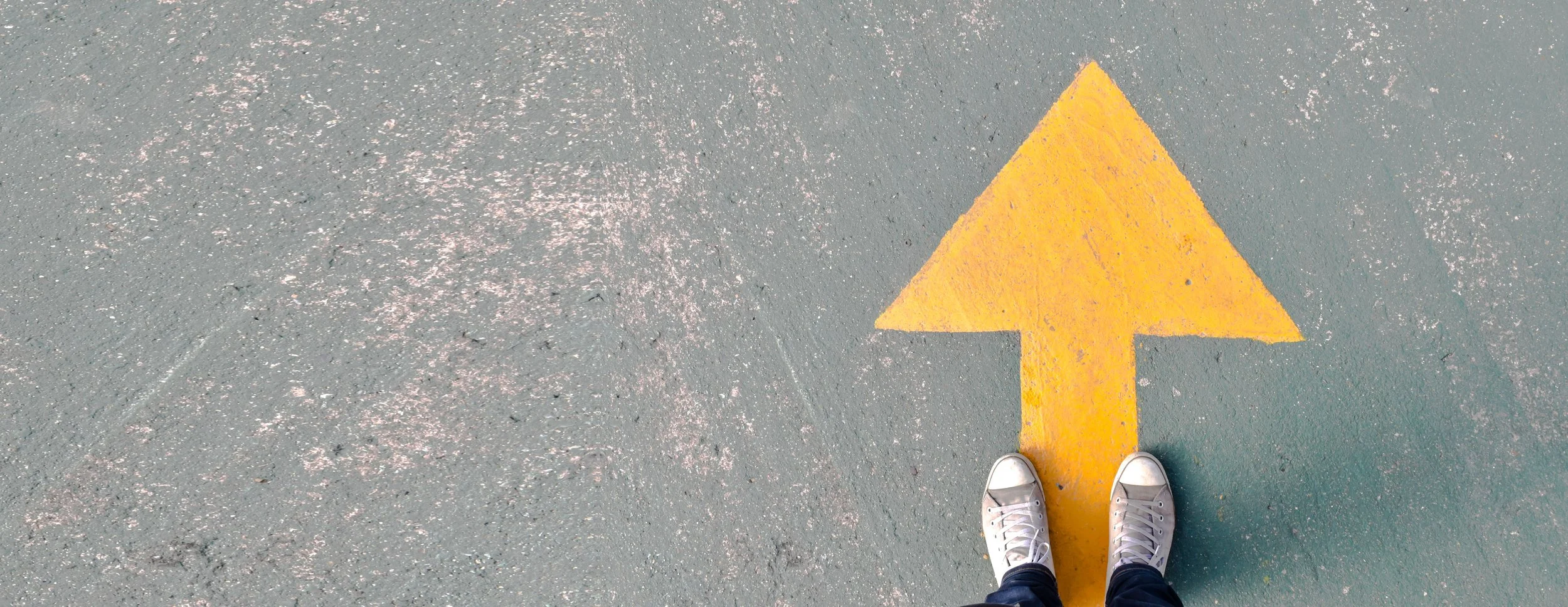 A student wearing white converse sneakers and black pants standing on a gray asphalt surface with a yellow upward arrow painted on it.