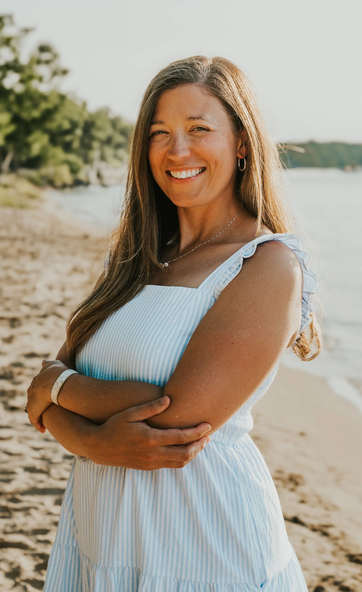 Smiling woman in a light blue and white striped dress standing on a beach with water and trees in the background.