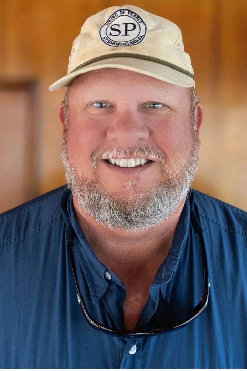 A smiling middle-aged man with a beard wearing a beige cap and sunglasses hanging from his shirt. He is dressed in a blue button-down shirt with a blurred warm-toned background.