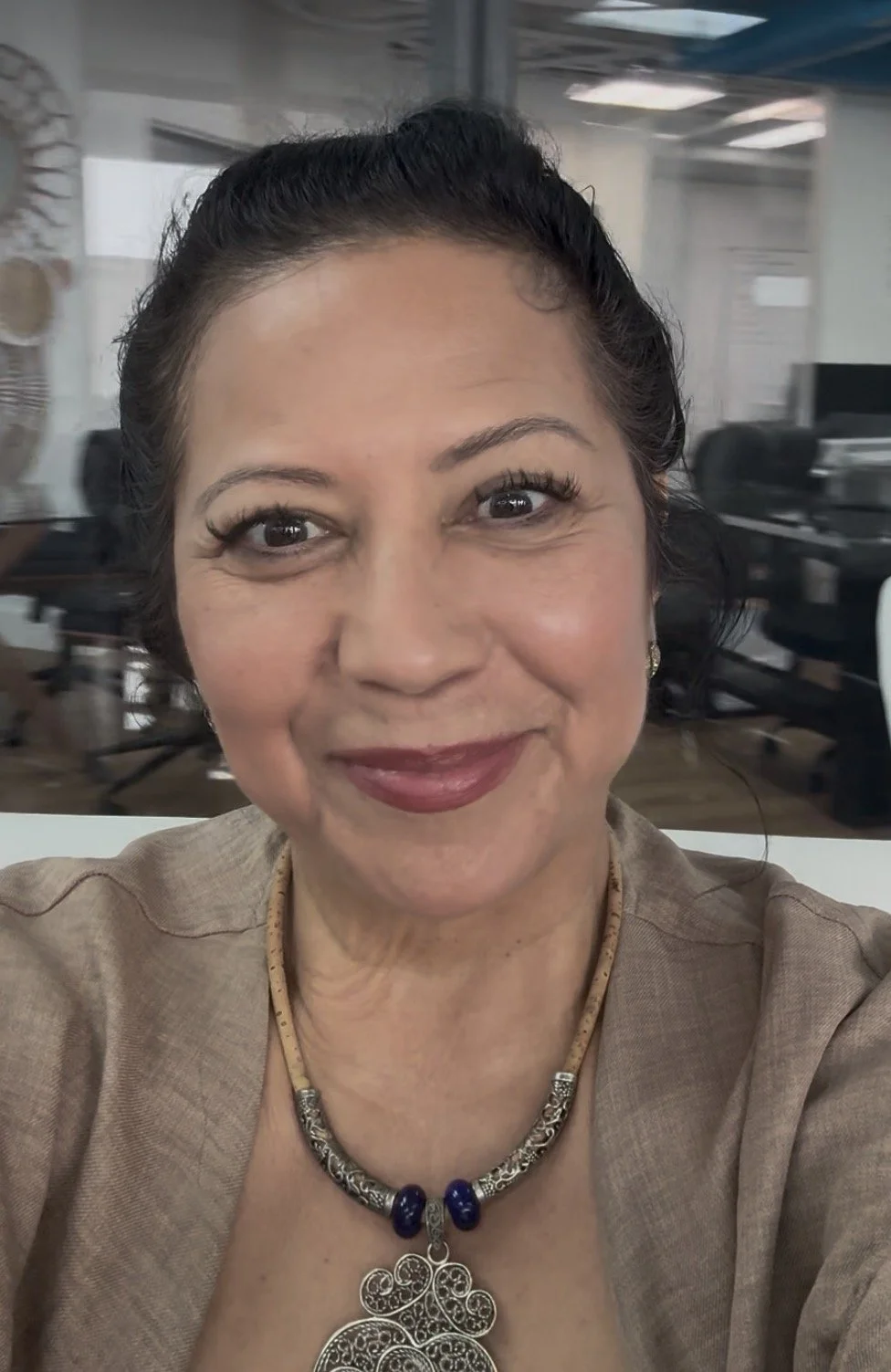 A woman with dark hair styled in an updo, wearing makeup, a beige blazer, and ornate jewelry, smiling at the camera in what appears to be an office environment.