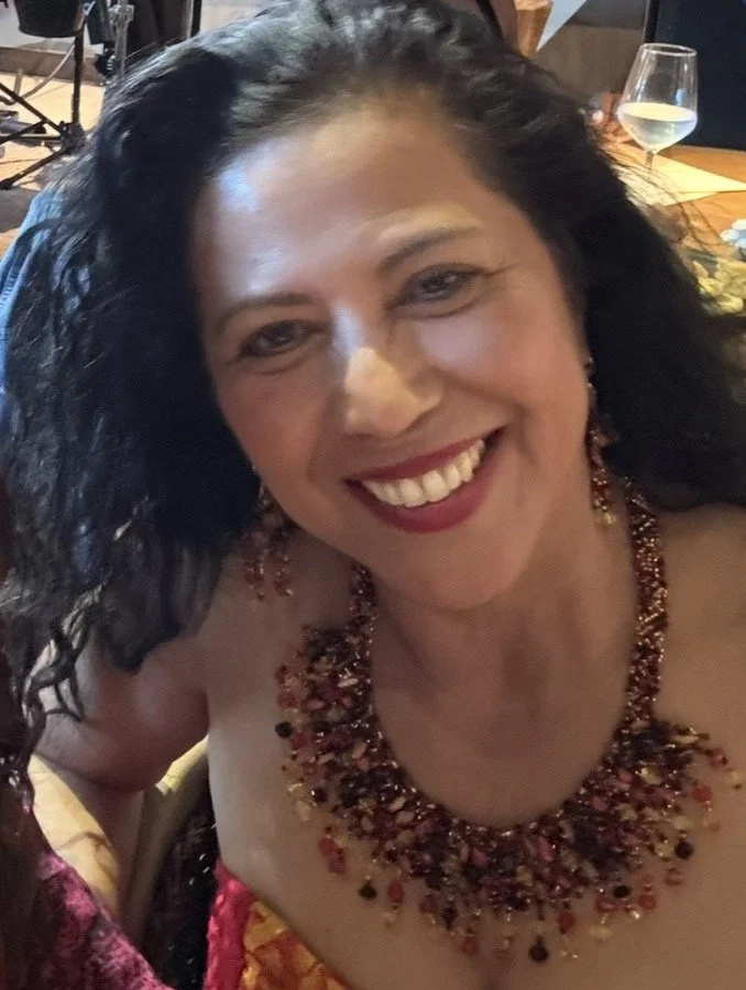 A smiling woman with curly black hair, wearing a red and gold beaded necklace and earrings, at a social gathering.
