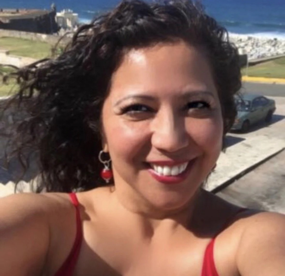 A woman smiling outdoors near the coast with buildings,-to the sea, and cars in the background.