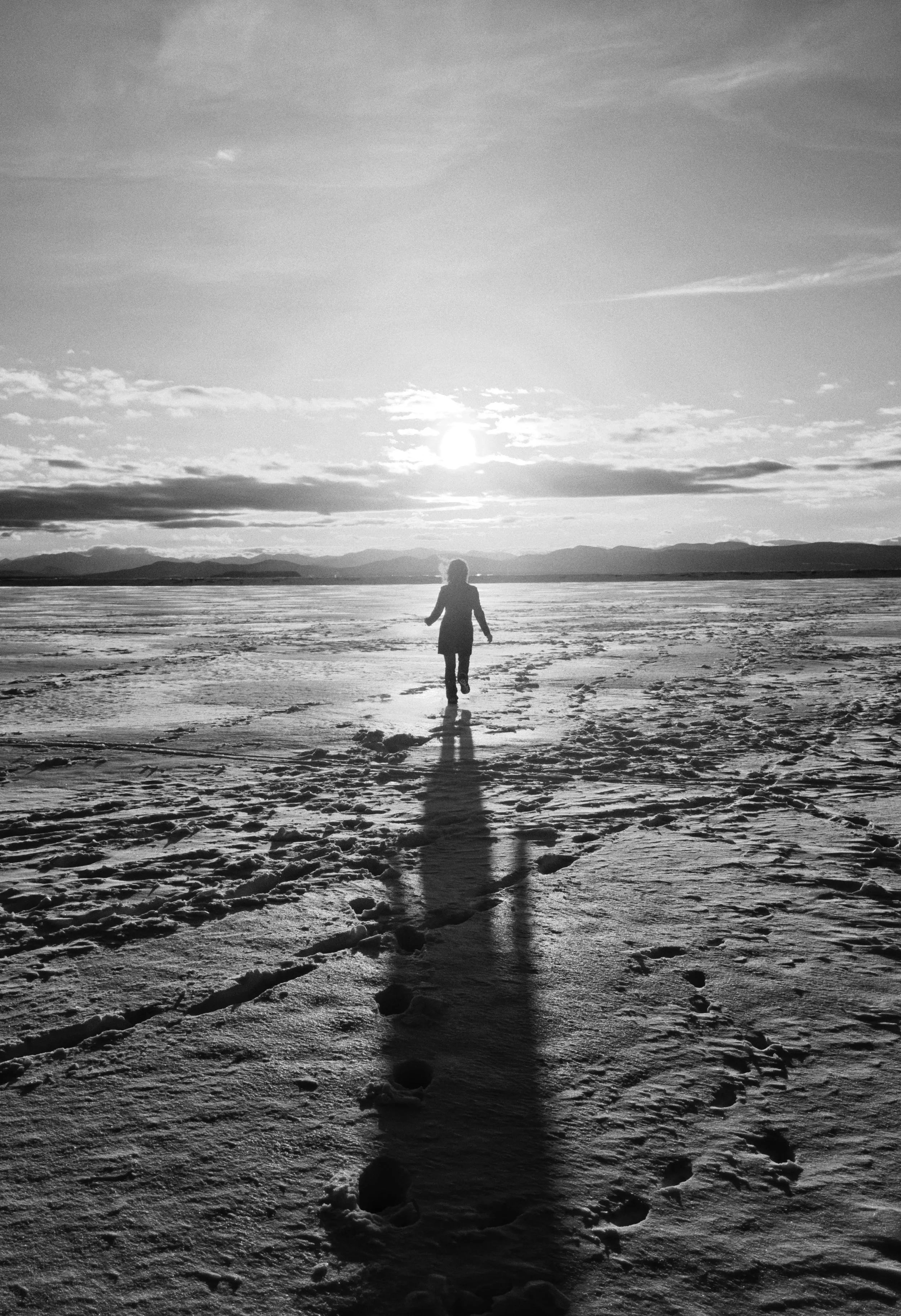 A person walking on a snowy landscape at sunset or sunrise, with mountains in the background and clouds in the sky.