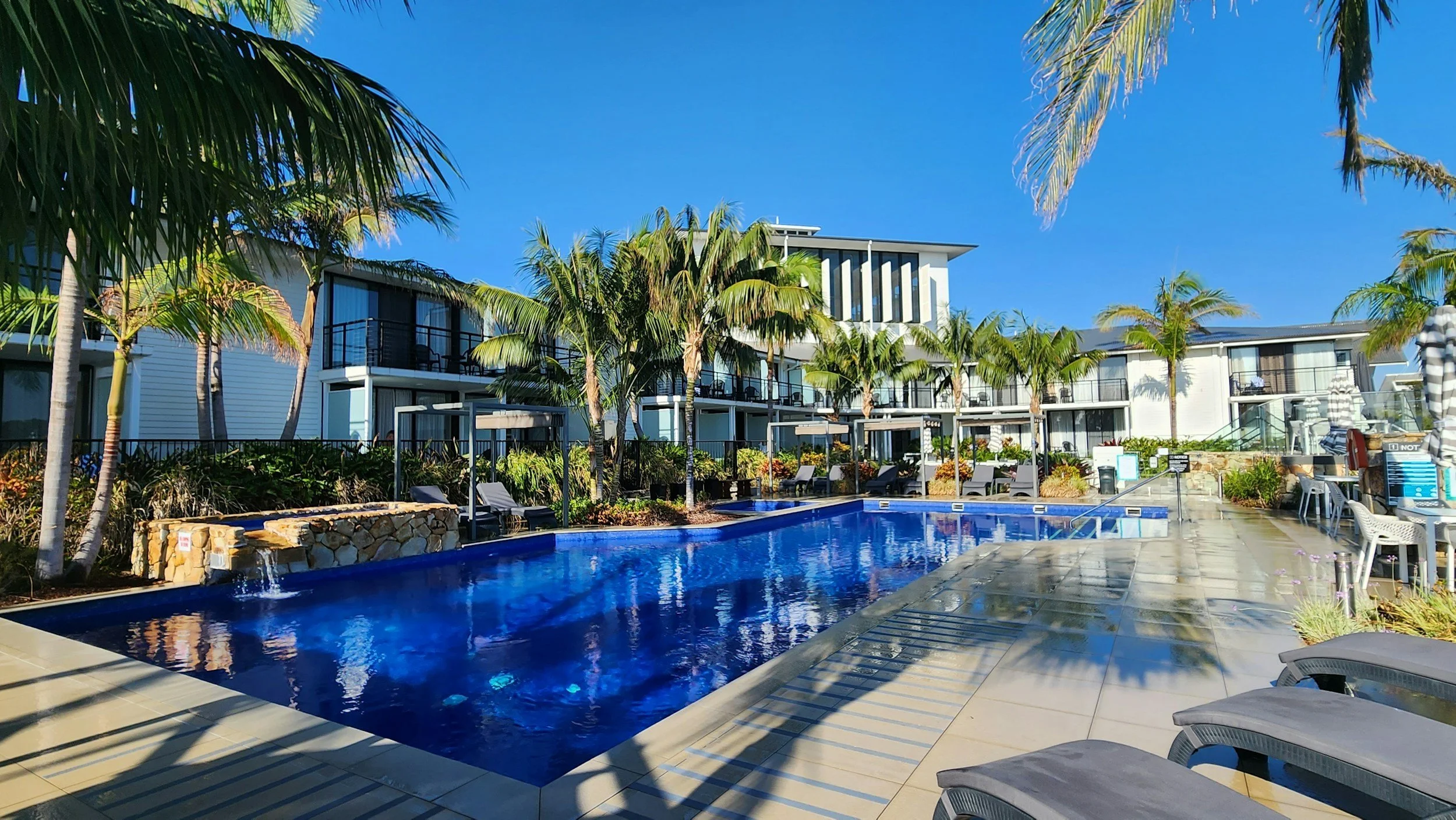 A resort swimming pool area surrounded by palm trees and modern white accommodations with balconies, with clear blue sky overhead.