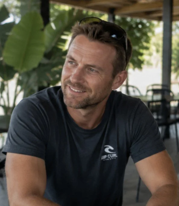 A man with short brown hair and light facial hair, wearing a dark gray T-shirt with a small logo, sitting outdoors at a table, smiling slightly, with large green leaves and a shaded patio area in the background.