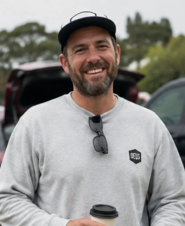 A smiling man outdoors wearing a gray sweatshirt, a black cap, and sunglasses hanging from his shirt collar. He is holding a coffee cup and standing in front of parked cars with trees in the background.