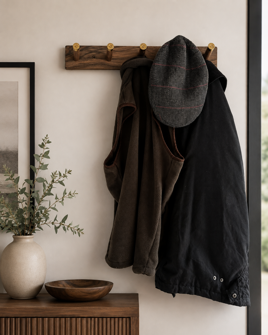 A wooden coat rack with three hooks holding a black jacket, a brown vest, and a gray plaid cap. There is a framed picture and a potted plant on a wooden table below the coat rack.