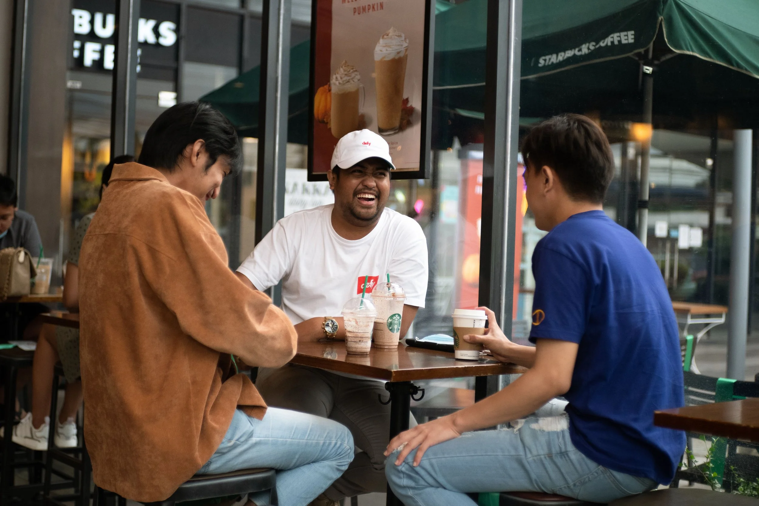 A group of friends enjoying drinks at an outdoor Starbucks, sitting at a high table with two frappuccinos and a coffee cup among them, engaged in conversation and laughing.