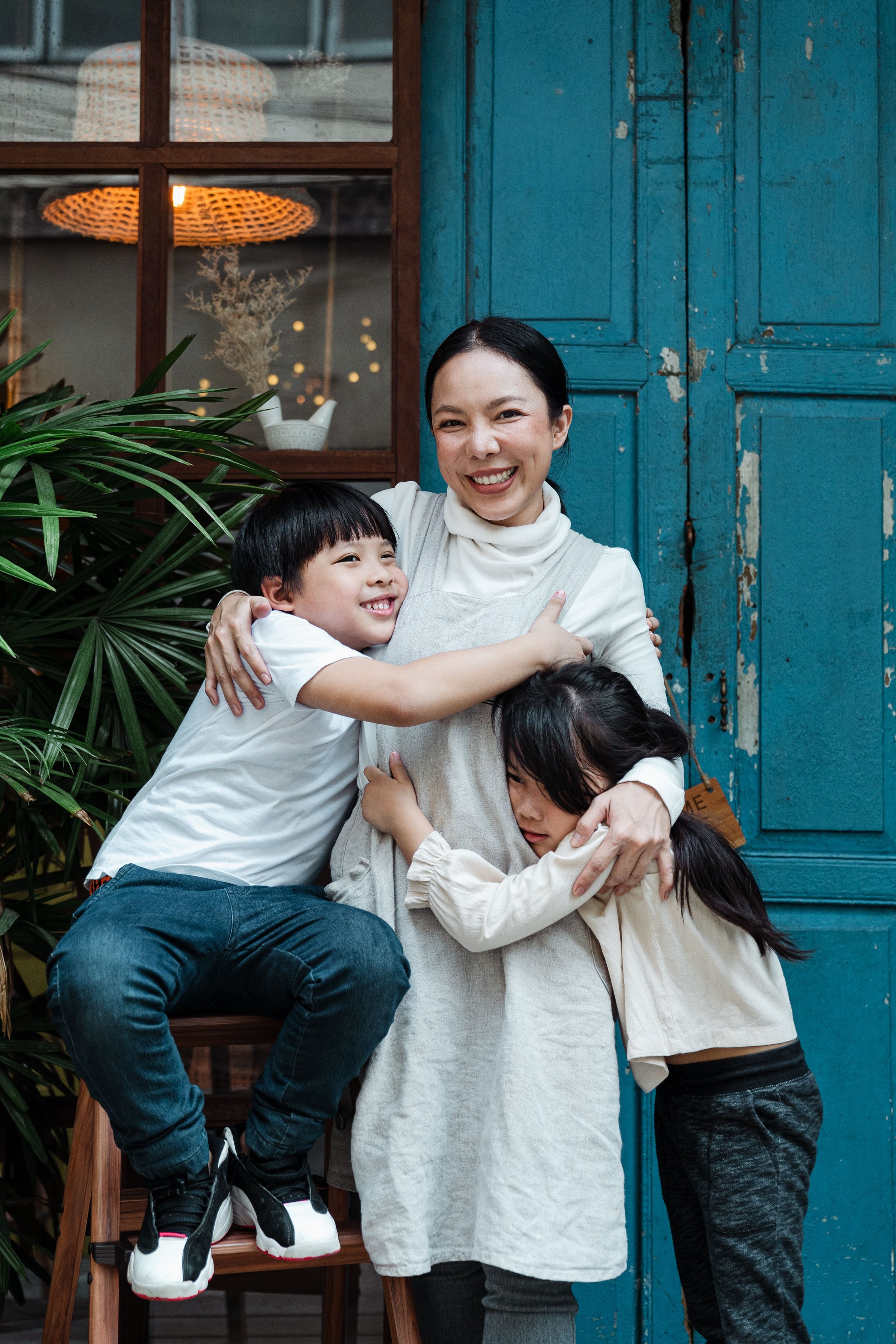 A woman with dark hair smiling and hugging three children in front of a blue wooden door with a window and a plant to the side.