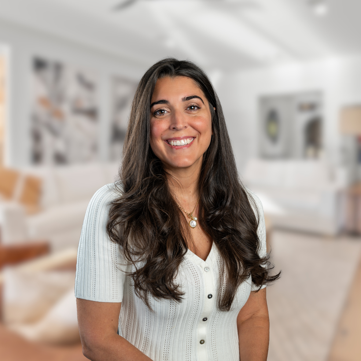 A smiling woman with long dark hair wearing a white button-up top and a gold necklace, standing in a blurred modern living room background.