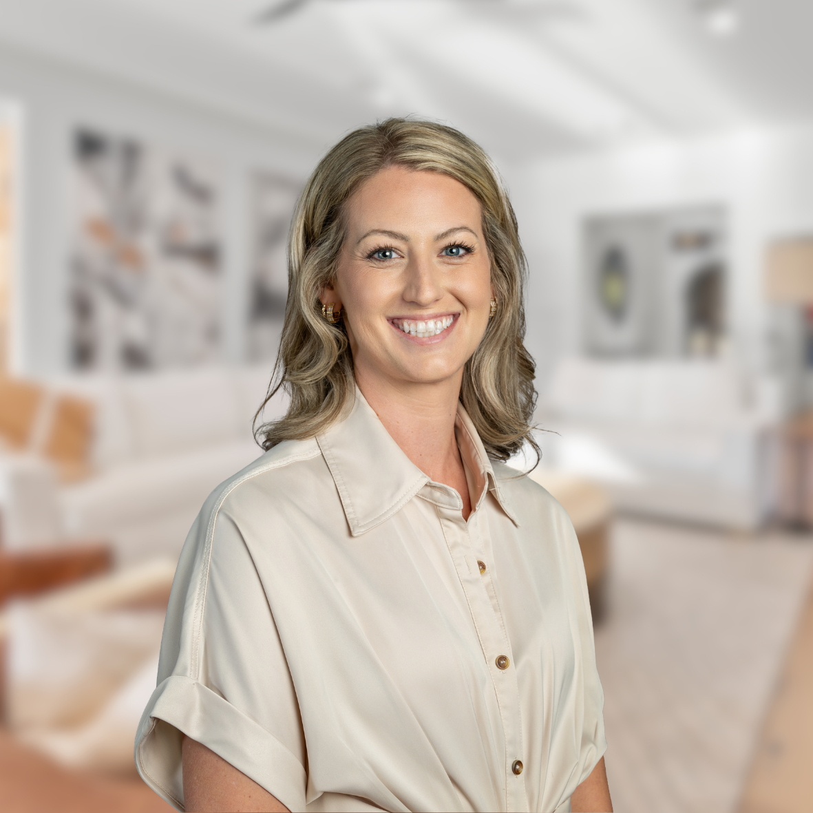 A smiling woman with blonde hair wearing a cream blouse in a blurred modern living room.