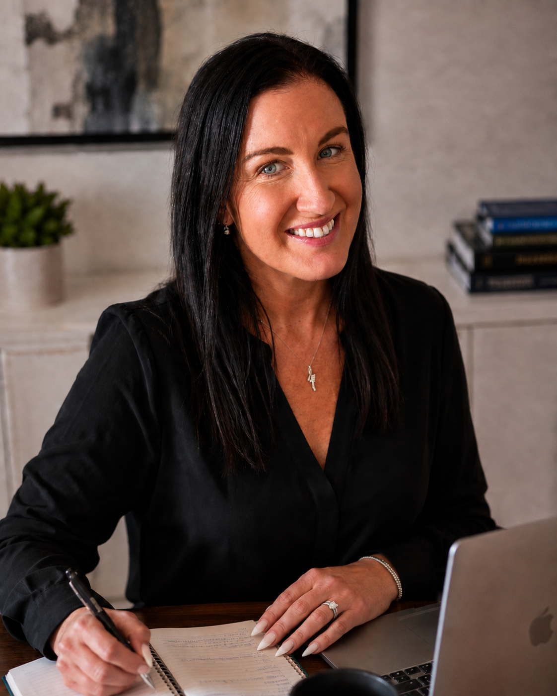 A woman with black hair and blue eyes smiling while sitting at a desk with a laptop, notebook, and pen in a well-lit office space.