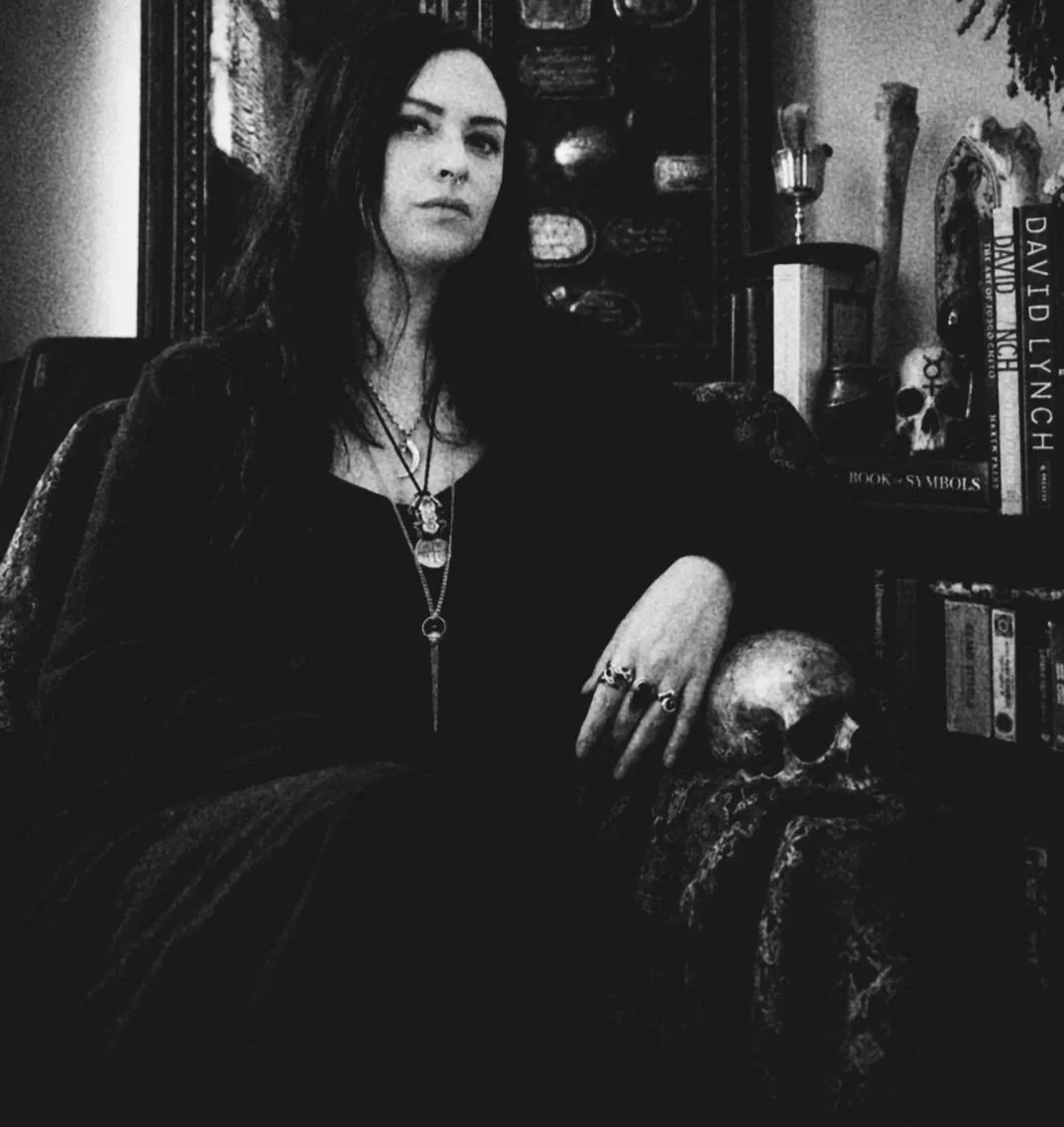 A woman with long dark hair sits beside a skull on a table in a dimly lit room, with books and mystical objects on shelves behind her.