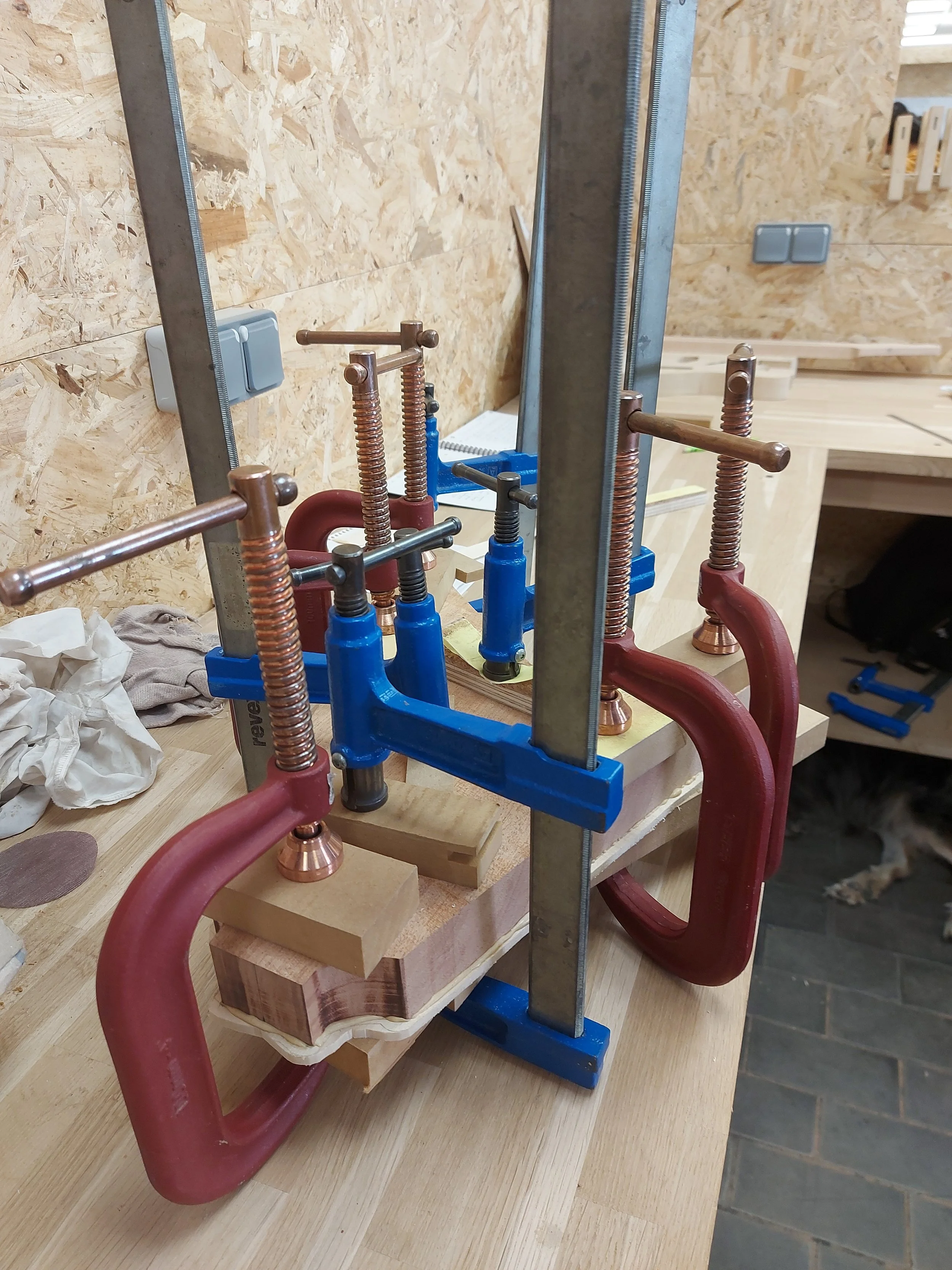 Un assemblage de pièces en cuivre et en acier maintenues par des serre-joints sur une table en bois, dans un environnement de travail ou d'atelier.