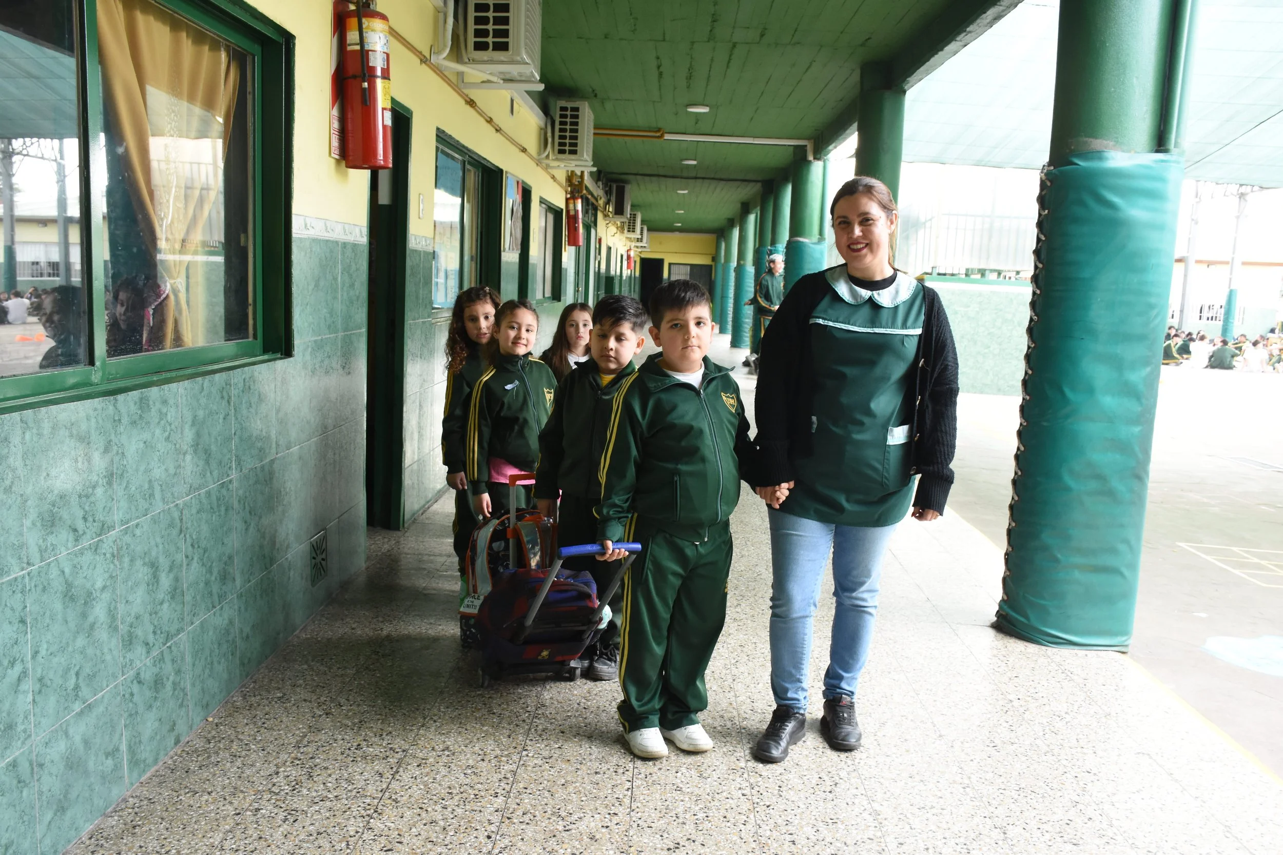 Una maestra sonriente camina junto a un grupo de estudiantes de primaria en fila por el pasillo del colegio, uno de ellos lleva un carrito de mochilas