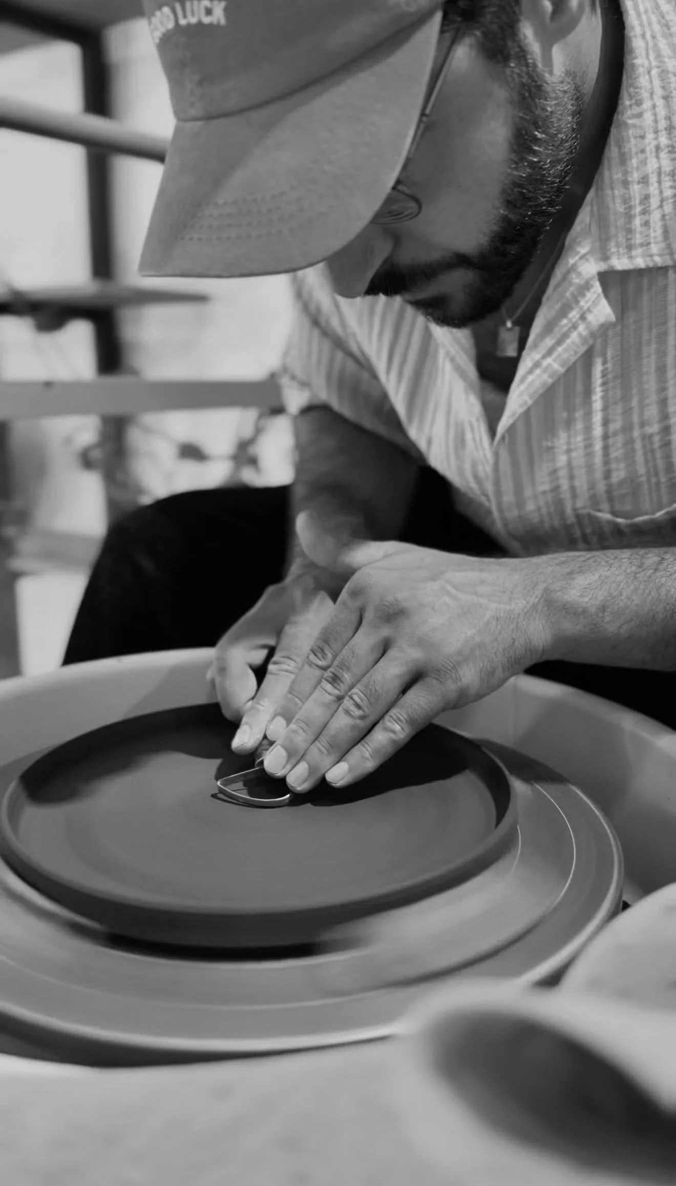 A man wearing glasses, a cap that says 'GOOD LUCK,' and a striped shirt, is working on a handmade ceramic plate in a studio setting.