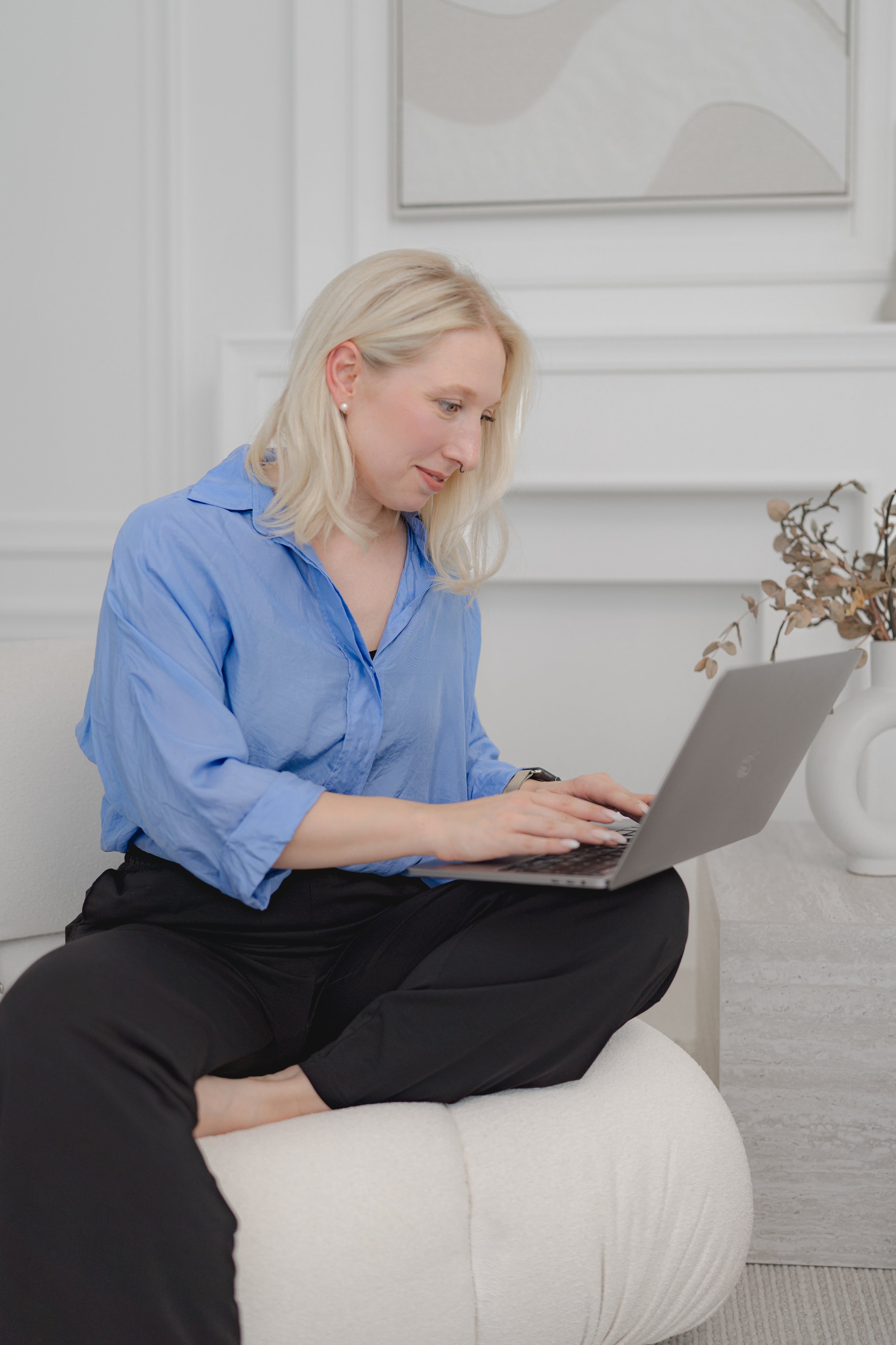 Eine blonde Frau in blauer Bluse sitzt auf einem weißen Sessel, arbeitet an einem Laptop und lächelt leicht.