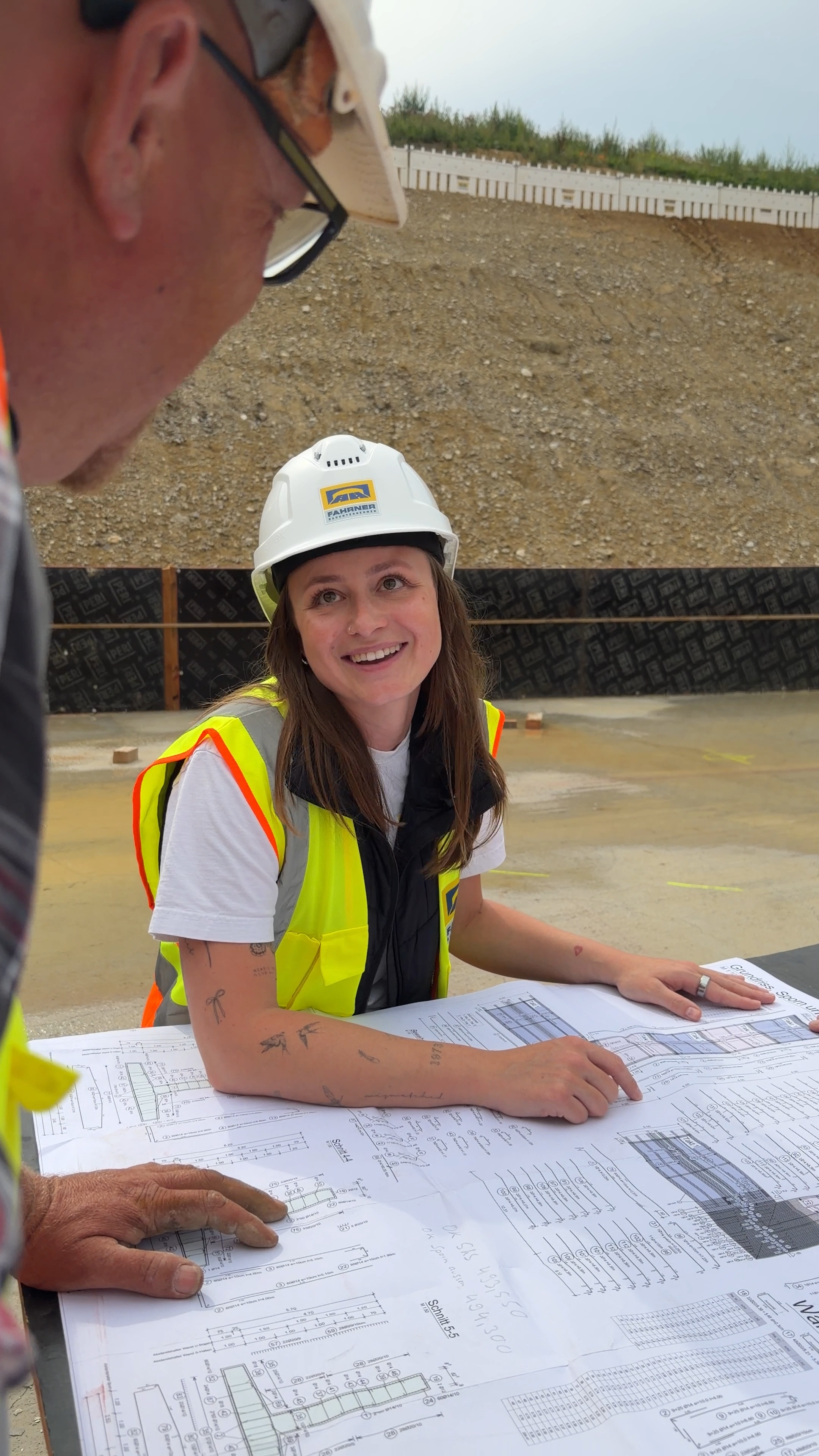 Beispiel für Employer Branding: Frau trägt einen weißen Bauhelm und eine gelbe Warnweste. Sie sitzt an einem Tisch mit Bauplänen und spricht wahrscheinlich mit einem Mann, der einen Bauhelm trägt. Baustelle im Hintergrund. Referenz Fahrner Bau