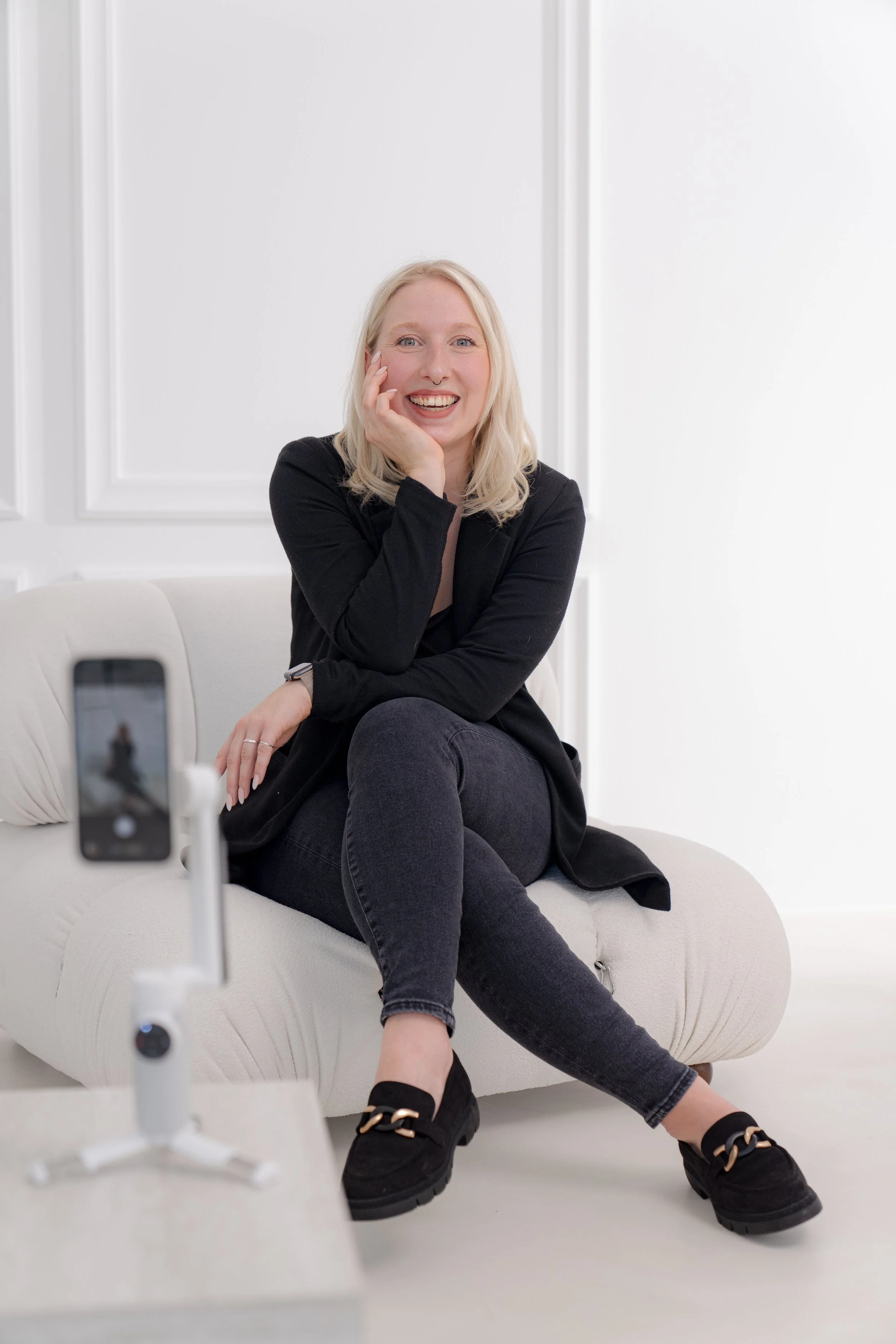 Lächelnde blonde Frau sitzt auf einer weißen Couch vor weißer Wand und macht ein Selfie mit einem Smartphone, das auf einem Stativ steht.