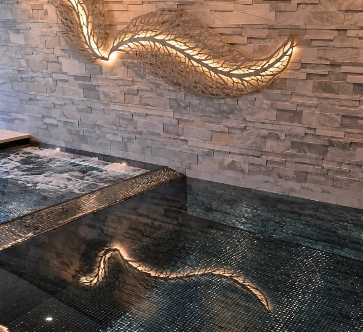 Bespoke illuminated leaf-inspired artwork on a natural stone feature wall beside the private hydropool, reflected in the dark blue water.