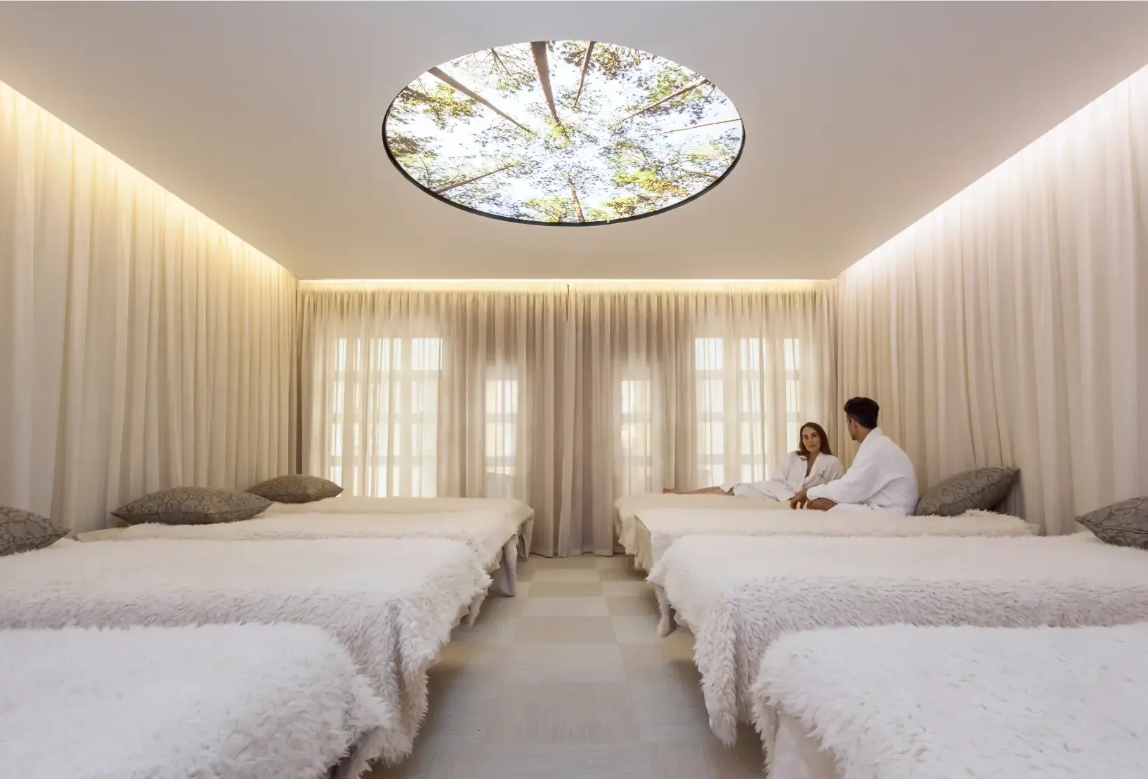 Two adults using the deep relax space at Champneys Forest Mere with sheer curtains and the bespoke ceiling light feature showing a view of trees and sky.