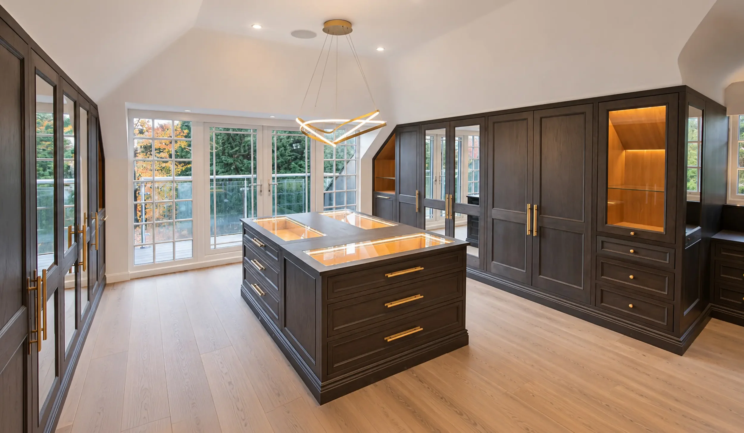 Bespoke dressing room in master bedroom in a luxury New Forest residence, featuring dark timber cabinetry, illuminated display shelving, central island and brass detailing.