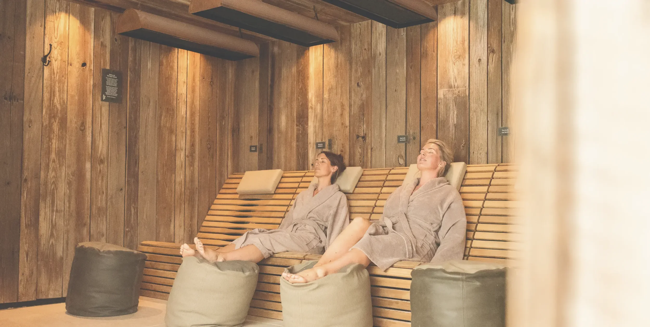 Two guests relaxing on heated wooden loungers in a warm, timber-clad spa relaxation room, designed for rest and recovery.