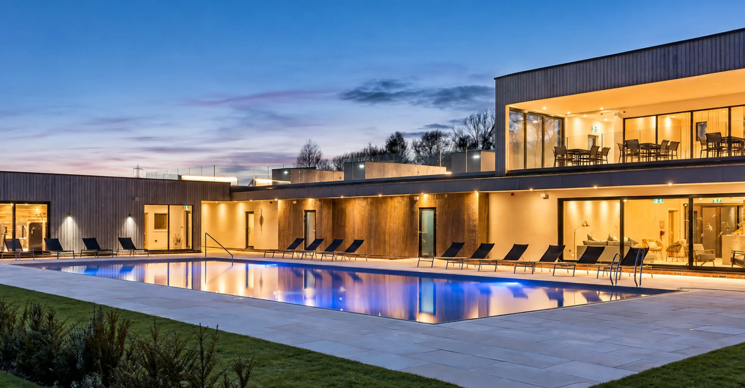 Lower Mill outdoor swimming pool at dusk with large glass windows revealing the gym and lounge area overlooking the pool and lake views