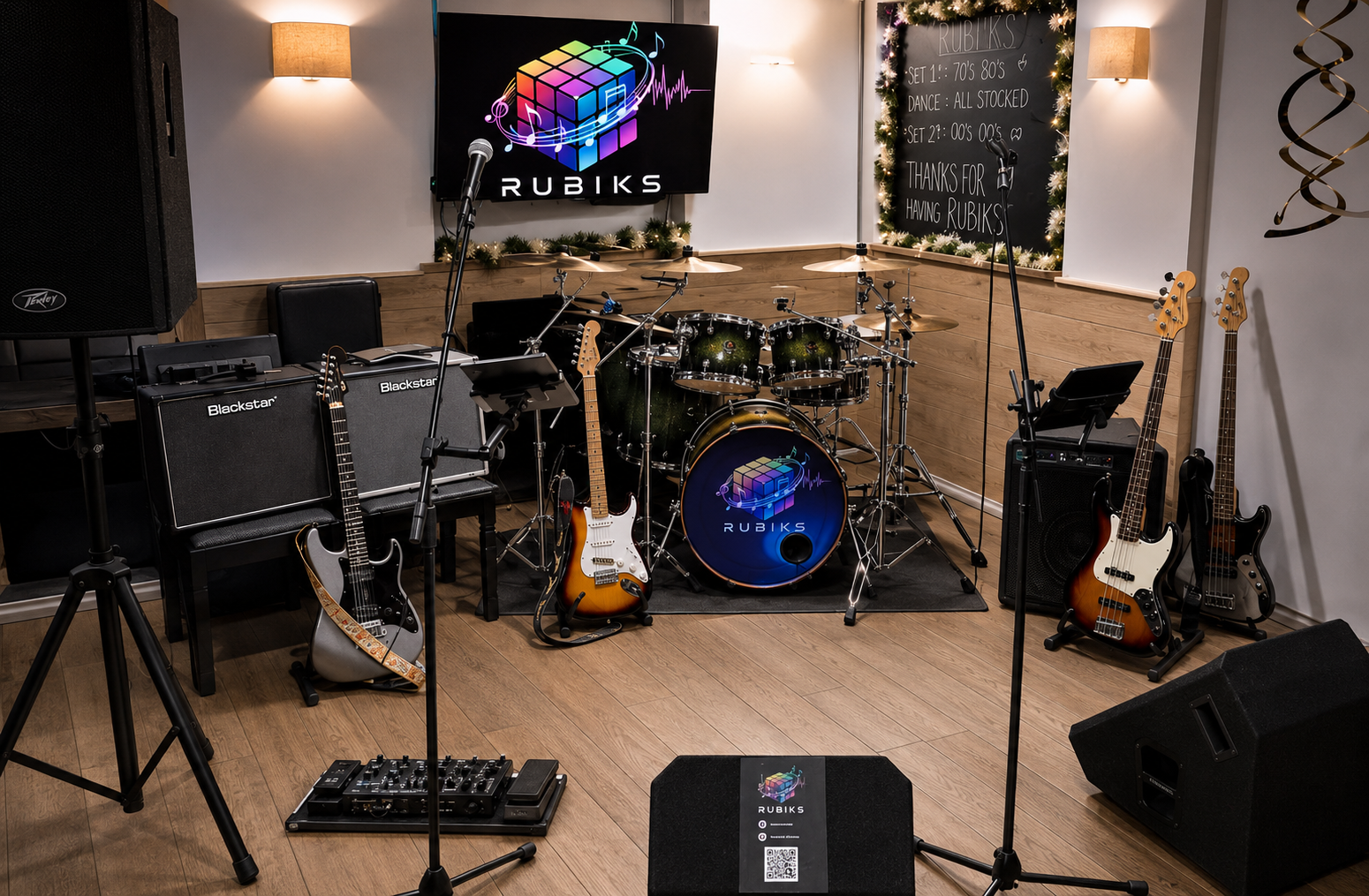 Music rehearsal room set up with drums, guitars, basses, amplifiers, microphone, and audio equipment, with a Rubiks logo and a board with song set lists.