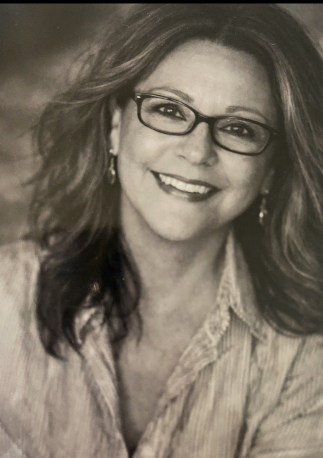 A black-and-white portrait of a smiling woman with glasses, earrings, and wavy hair, wearing a collared shirt.