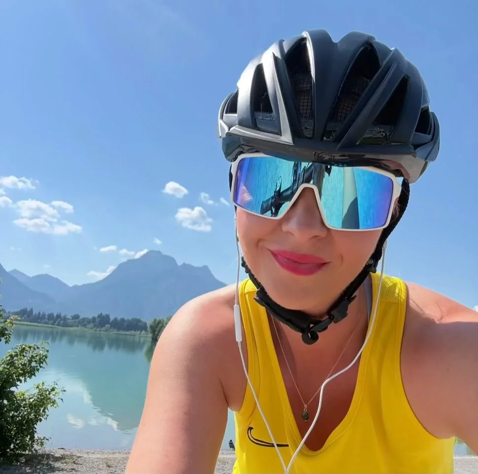 Lenka mit Fahrradhelm und gelbem Radtrikot am Forggensee mit Blick auf die Allgäuer Berge