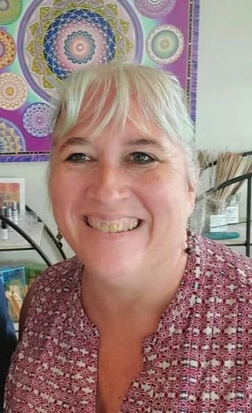 A smiling woman with blonde hair and wearing a red patterned top, standing in front of colorful mandala artwork on the wall, inside a room with shelves and art supplies.