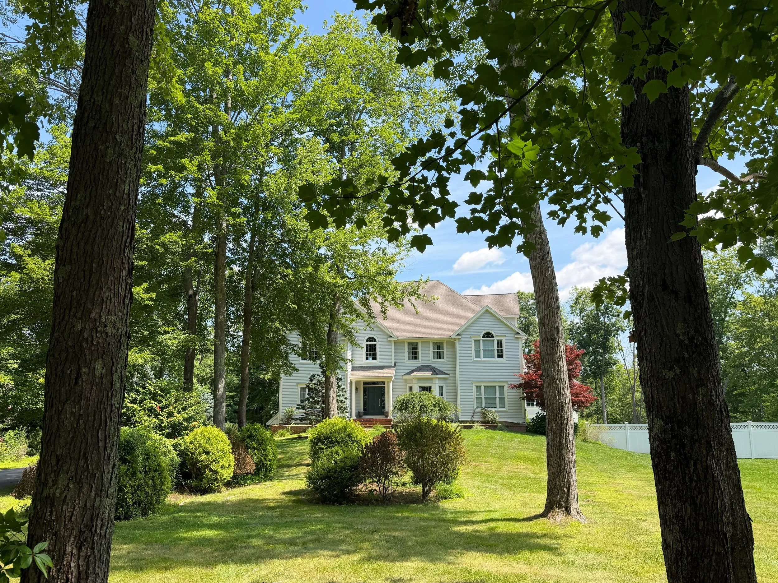 A white house with a triangular roof, multiple windows, and a front porch, framed by tall trees with lush green leaves and a well-manicured lawn.